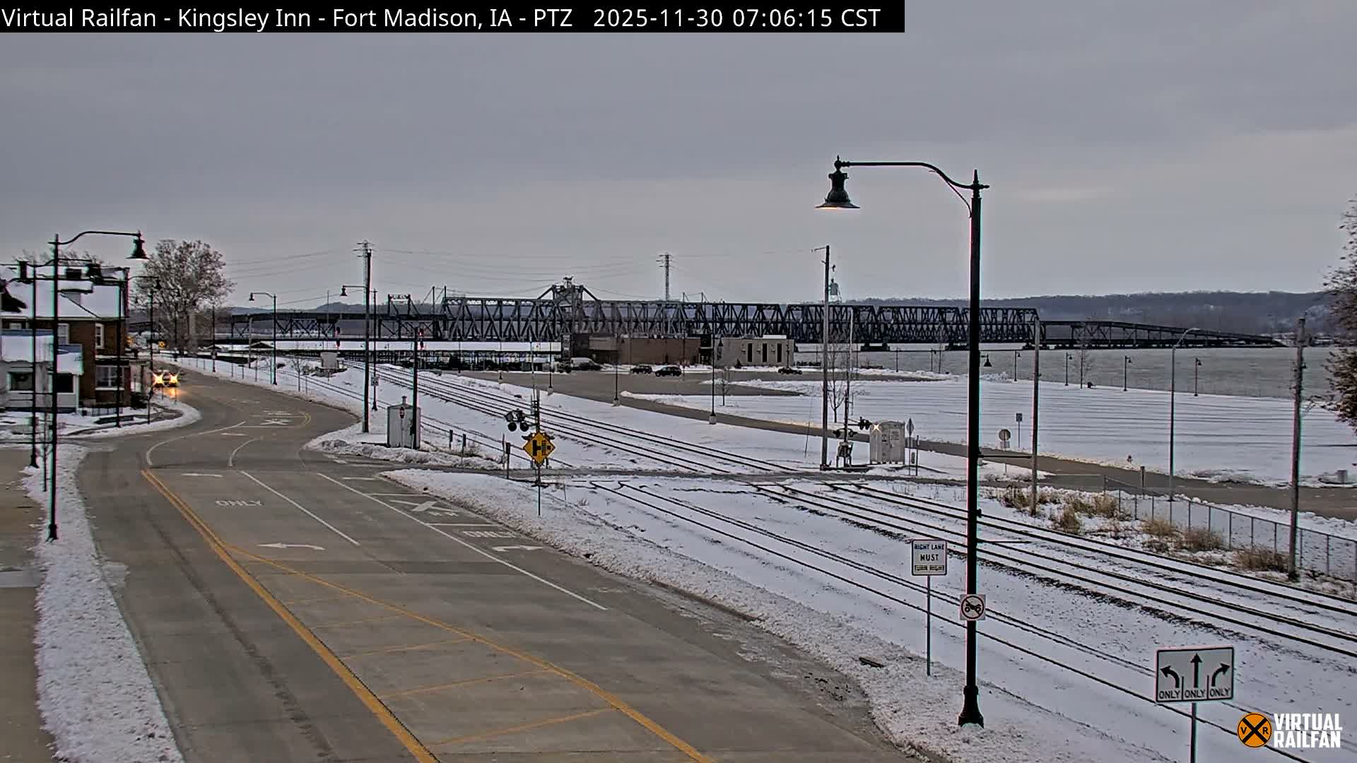 A clear night view displays a multi-lane road and multiple railroad tracks illuminated by numerous streetlights, with buildings and a large bridge visible in the distance.