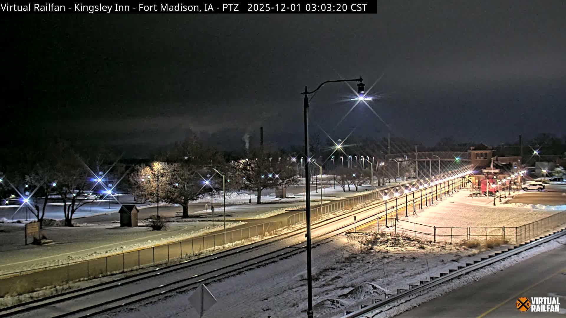 A snowy winter night scene depicts snow-covered train tracks, platforms, and bare trees illuminated by many streetlights, with a historic brick building and a red train car visible near the tracks under a dark, overcast sky.