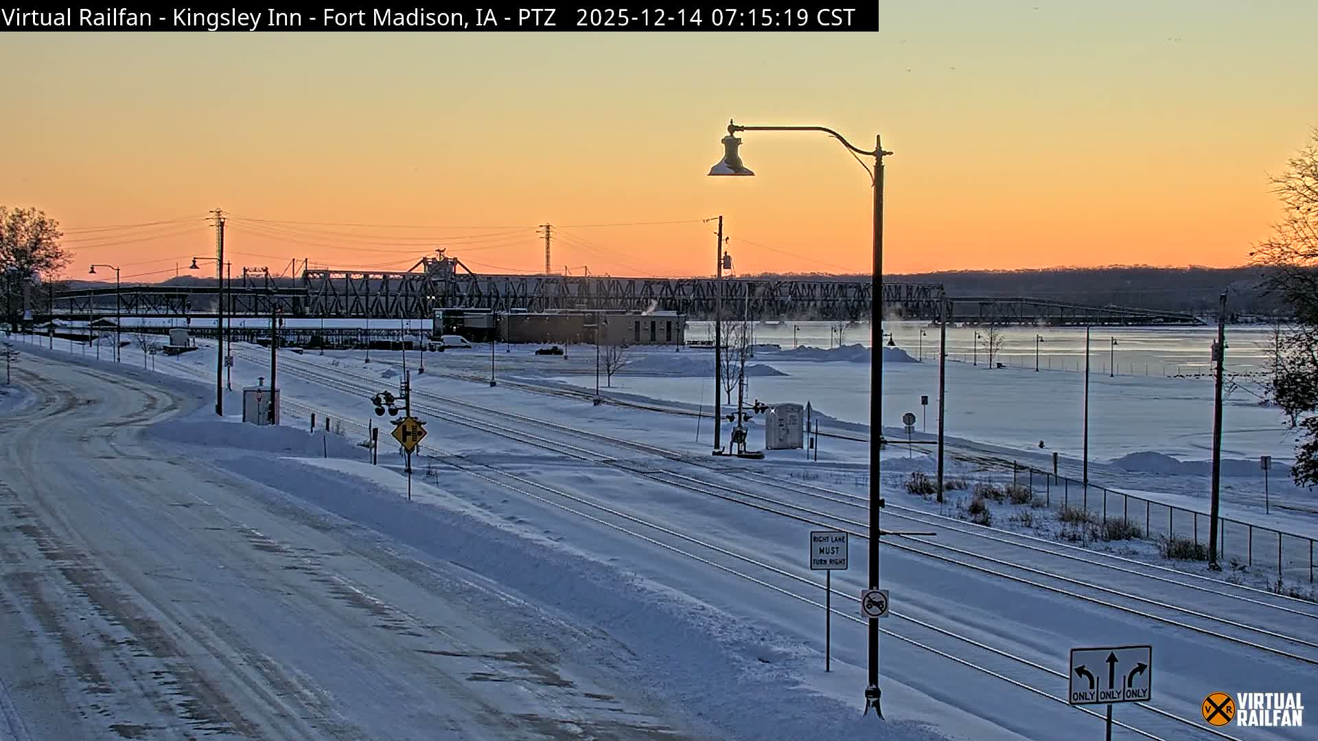 A snowy winter night scene depicts snow-covered train tracks, platforms, and bare trees illuminated by many streetlights, with a historic brick building and a red train car visible near the tracks under a dark, overcast sky.