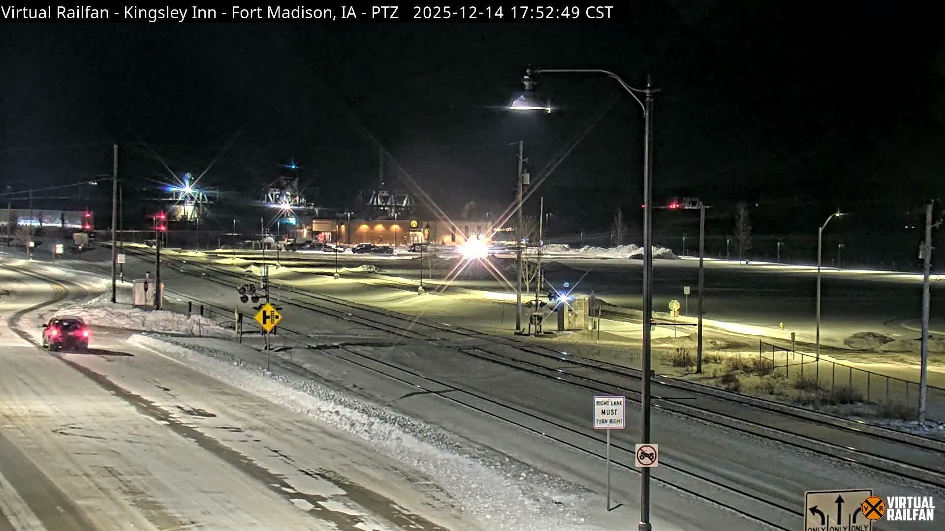 A snowy winter night scene depicts snow-covered train tracks, platforms, and bare trees illuminated by many streetlights, with a historic brick building and a red train car visible near the tracks under a dark, overcast sky.