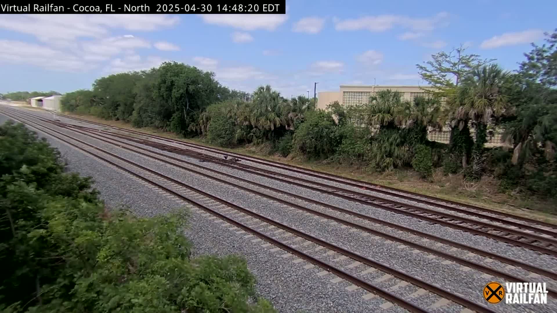 Multiple train tracks stretch into the distance under a partly cloudy sky, bordered by lush green vegetation and a building in the background.