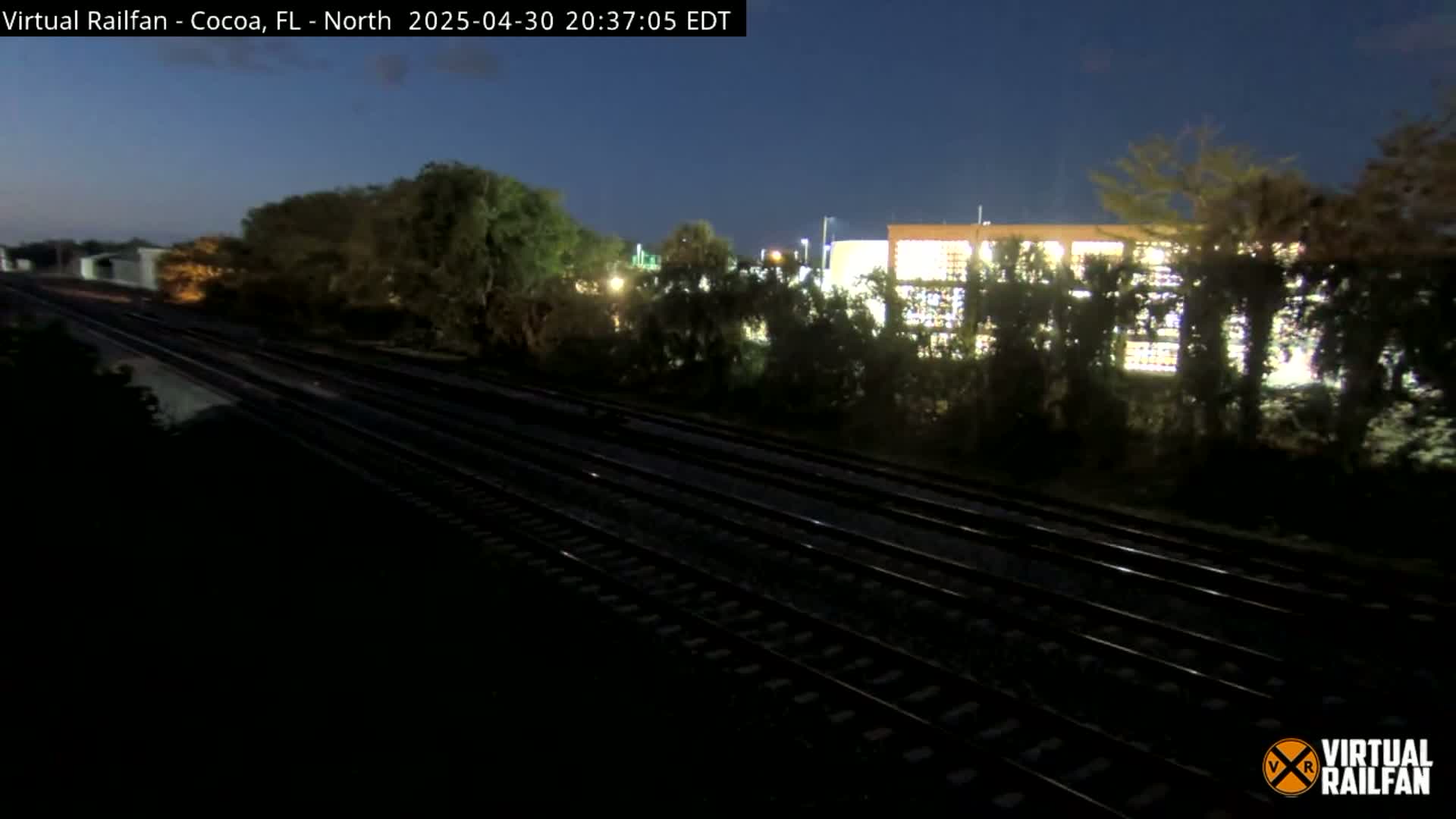 Multiple train tracks run through a darkly lit landscape at night, past vegetation and a brightly lit building.