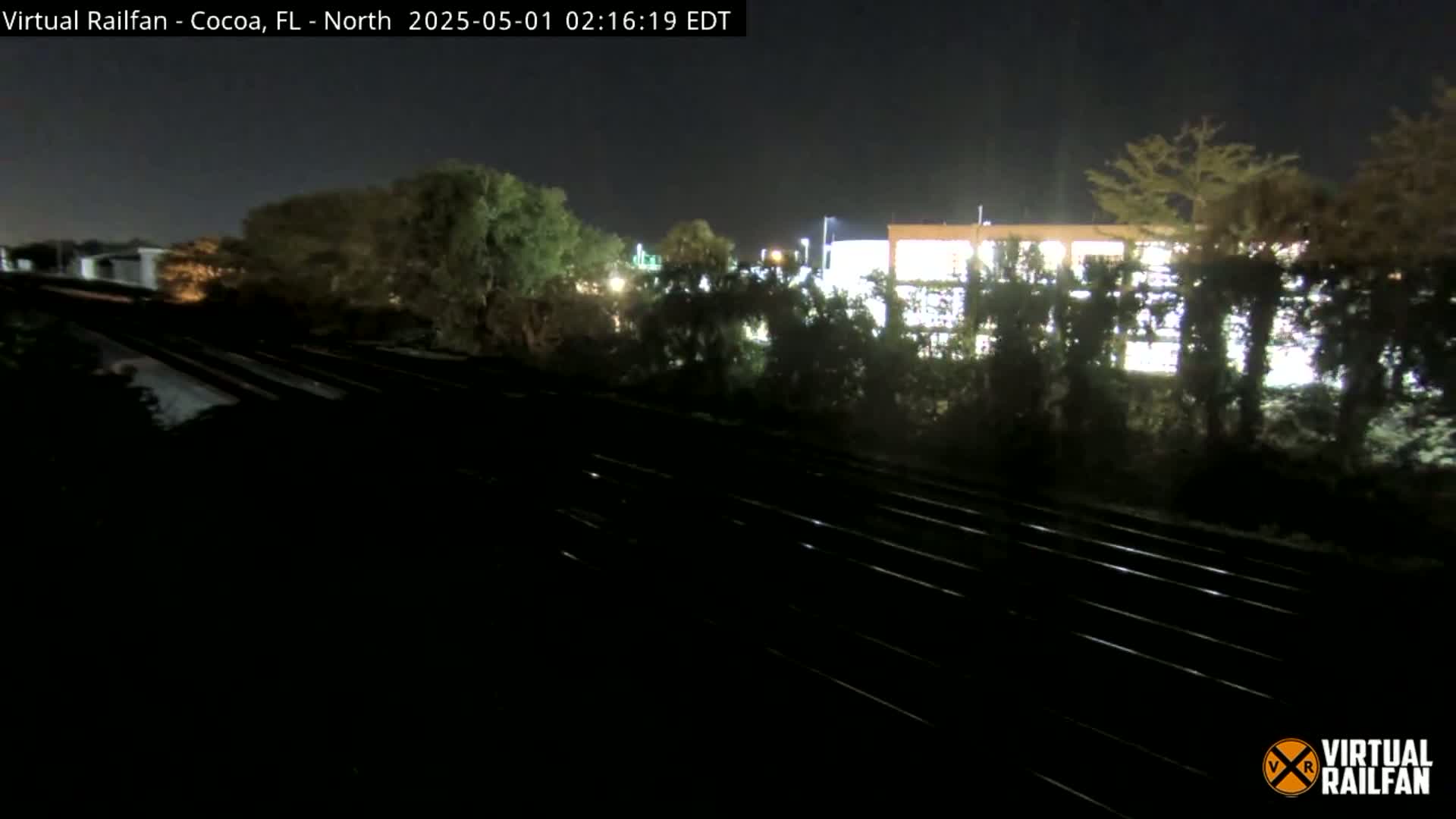 At night, a nighttime view shows train tracks extending into the distance past shrubbery and a building with lights on.