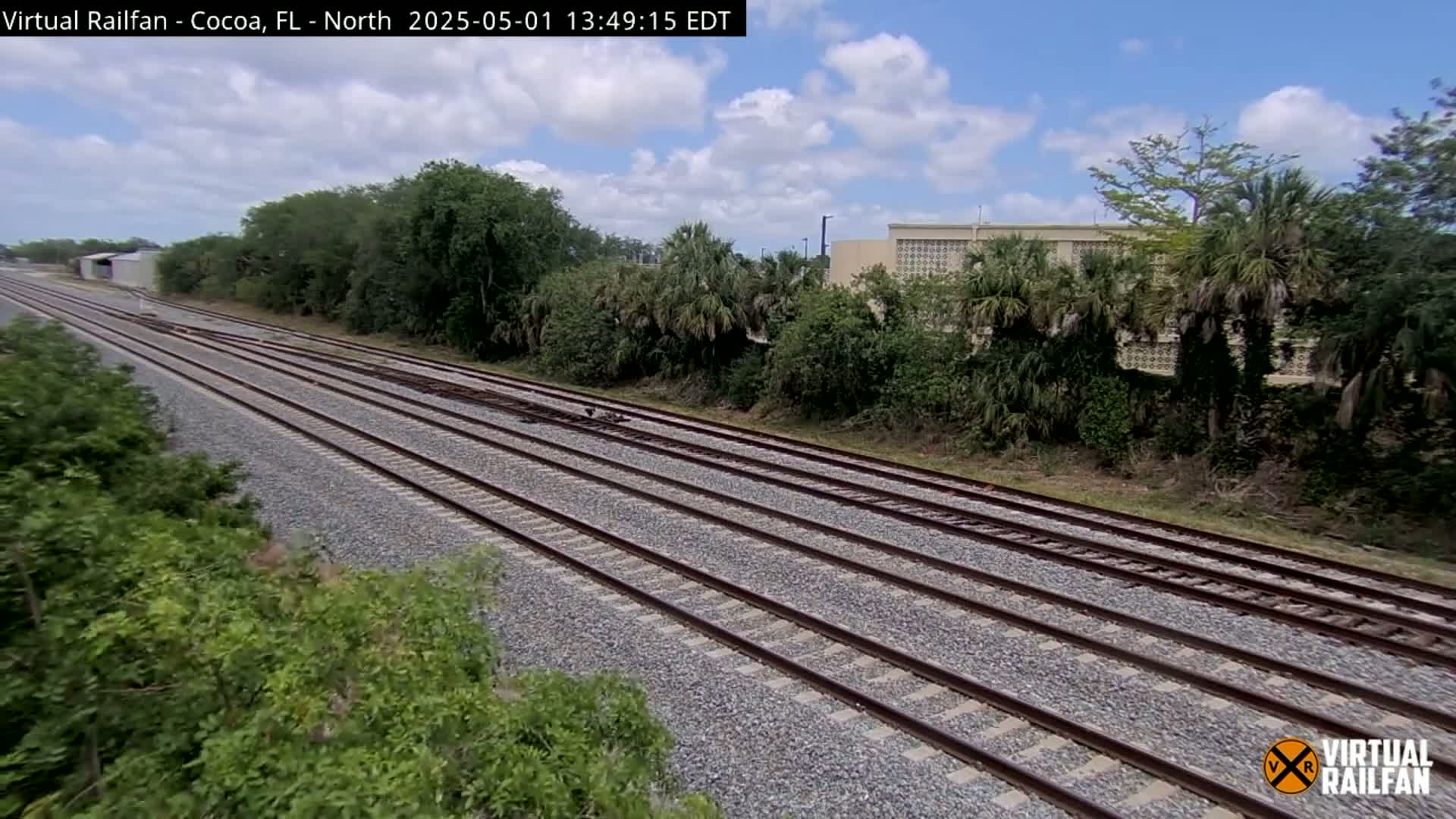 Multiple sets of train tracks stretch into the distance under a partly cloudy sky, bordered by lush green vegetation and a building.