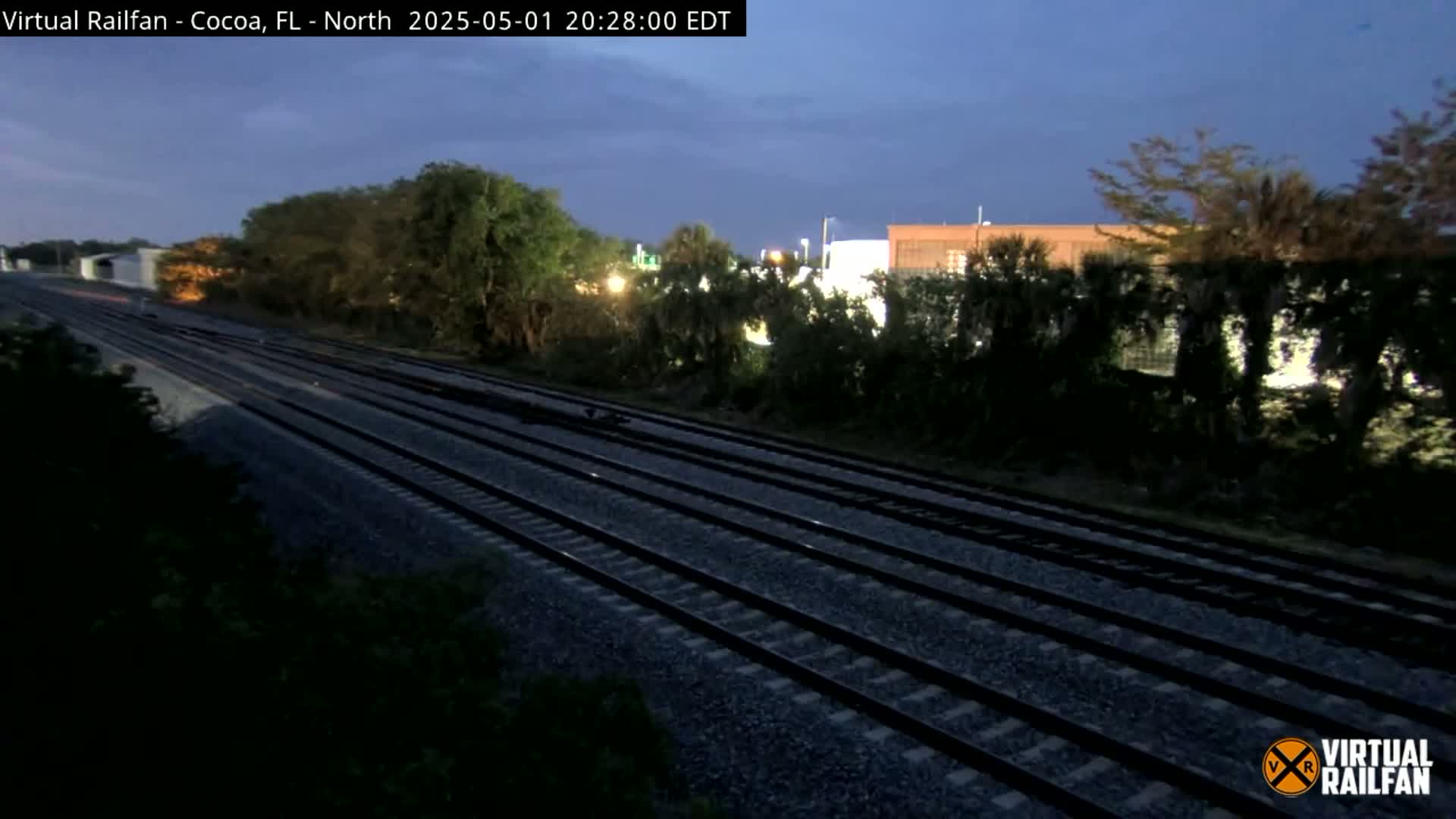 Multiple train tracks stretch into the distance under a twilight sky, bordered by dark green vegetation and a building in the background.