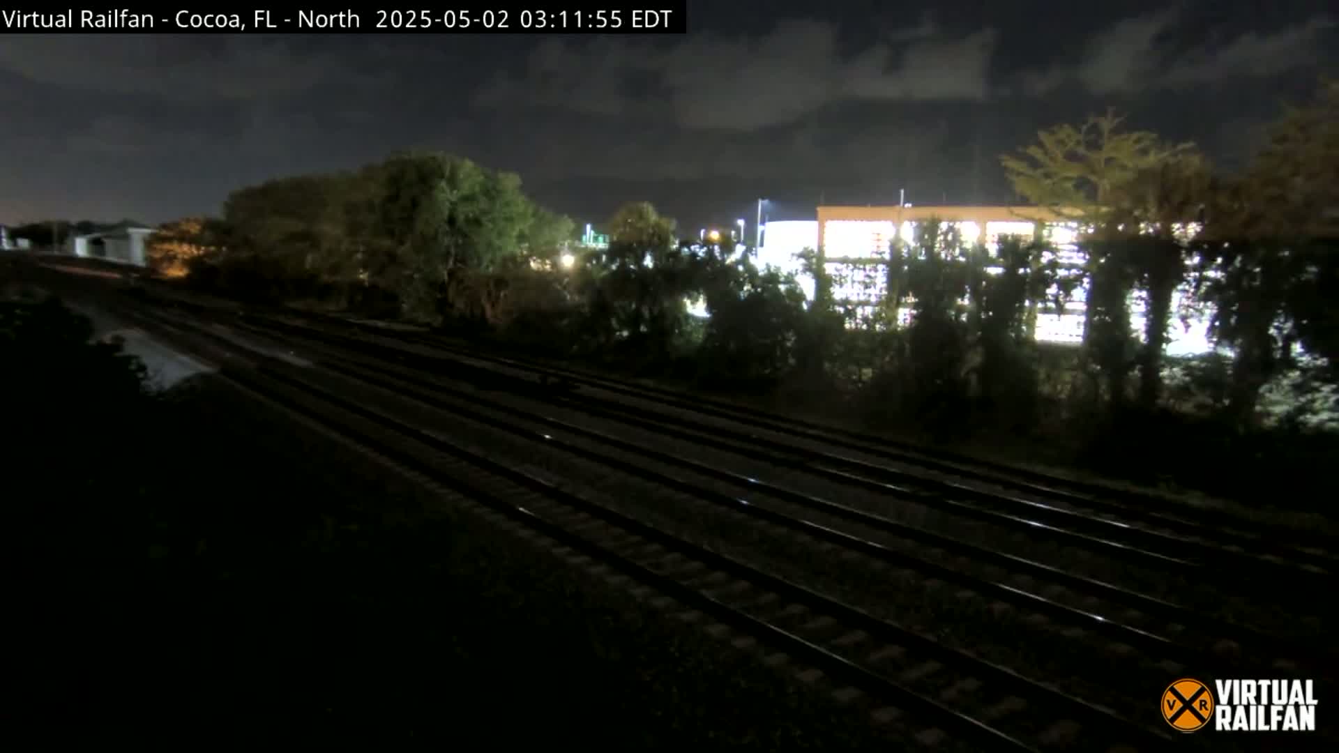 Multiple train tracks extend into the distance under a nighttime sky, past dark vegetation and a brightly lit building.