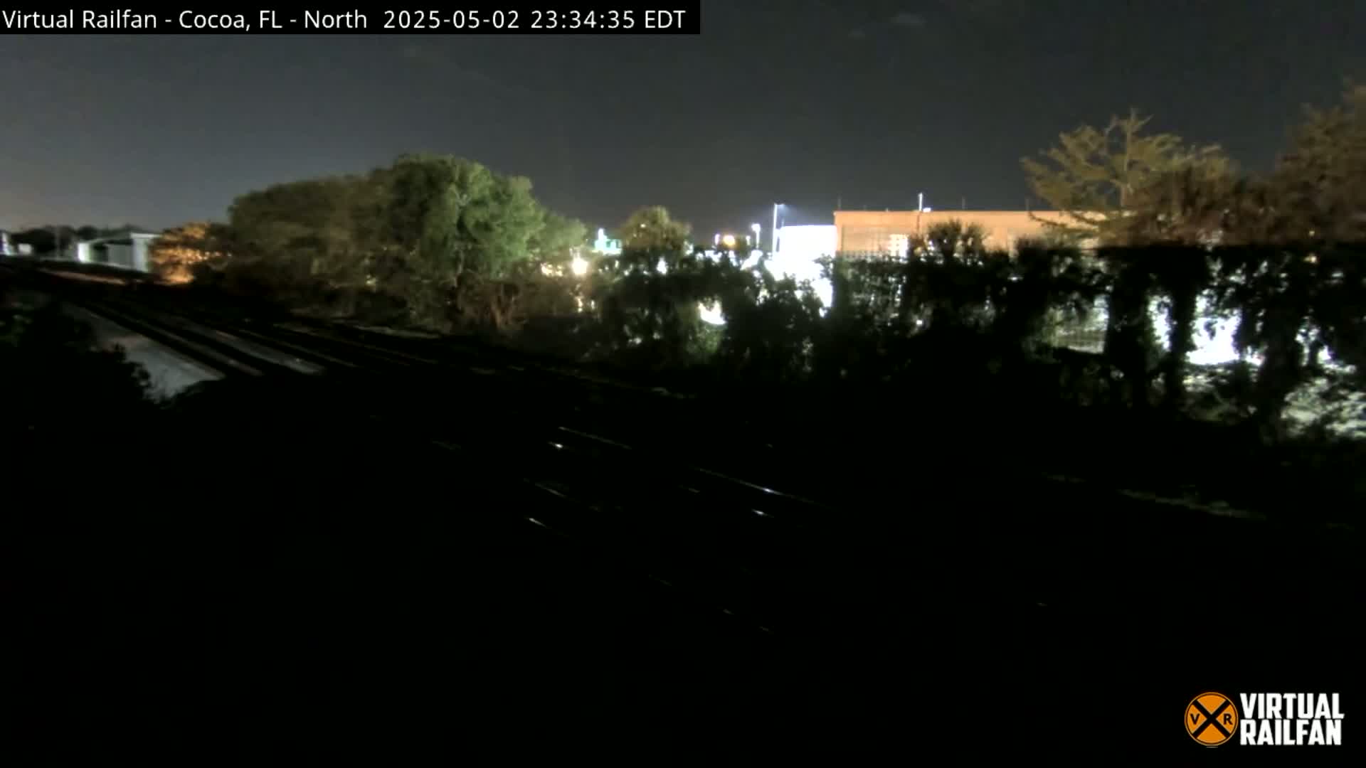 At night, train tracks run through a dark landscape of trees and shrubs, with a dimly lit building visible in the distance.