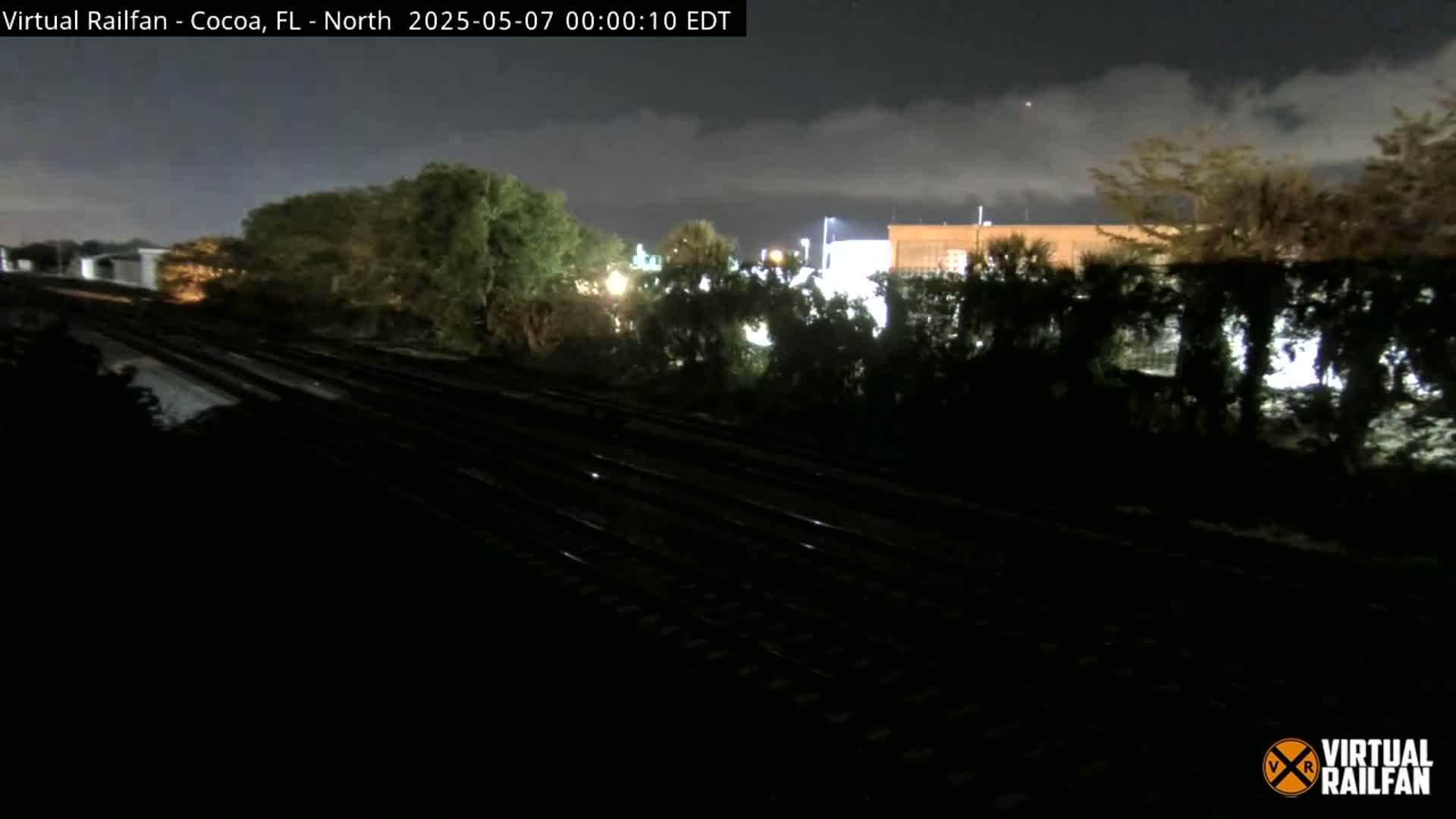 A nighttime view of train tracks running through a landscape of dark vegetation and buildings under a cloudy sky.