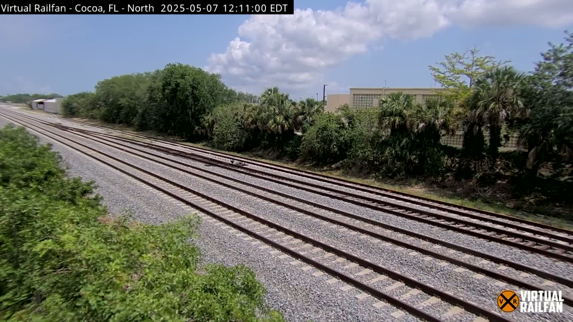 Multiple train tracks extend into the distance under a partly cloudy sky, bordered by lush green vegetation and a building in the background.