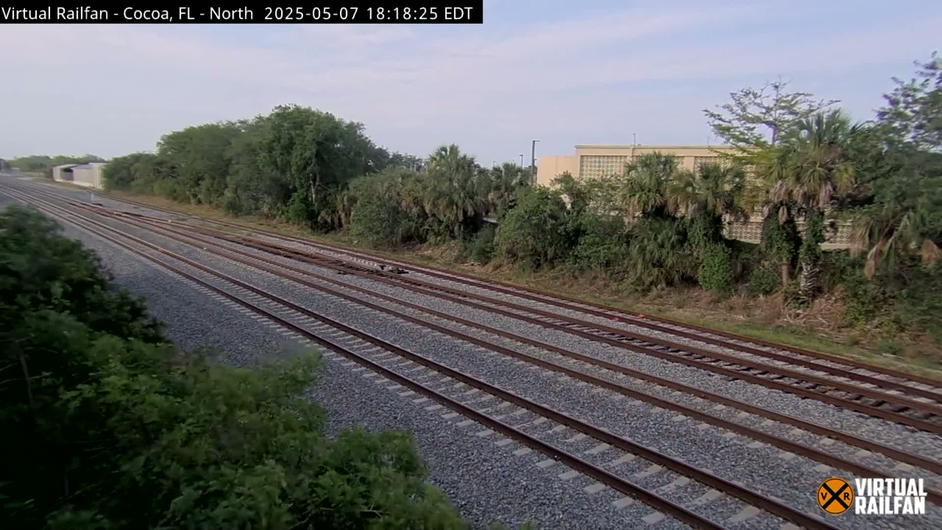 Multiple parallel train tracks extend into the distance under a partly cloudy sky, bordered by lush green vegetation and a building in the background.