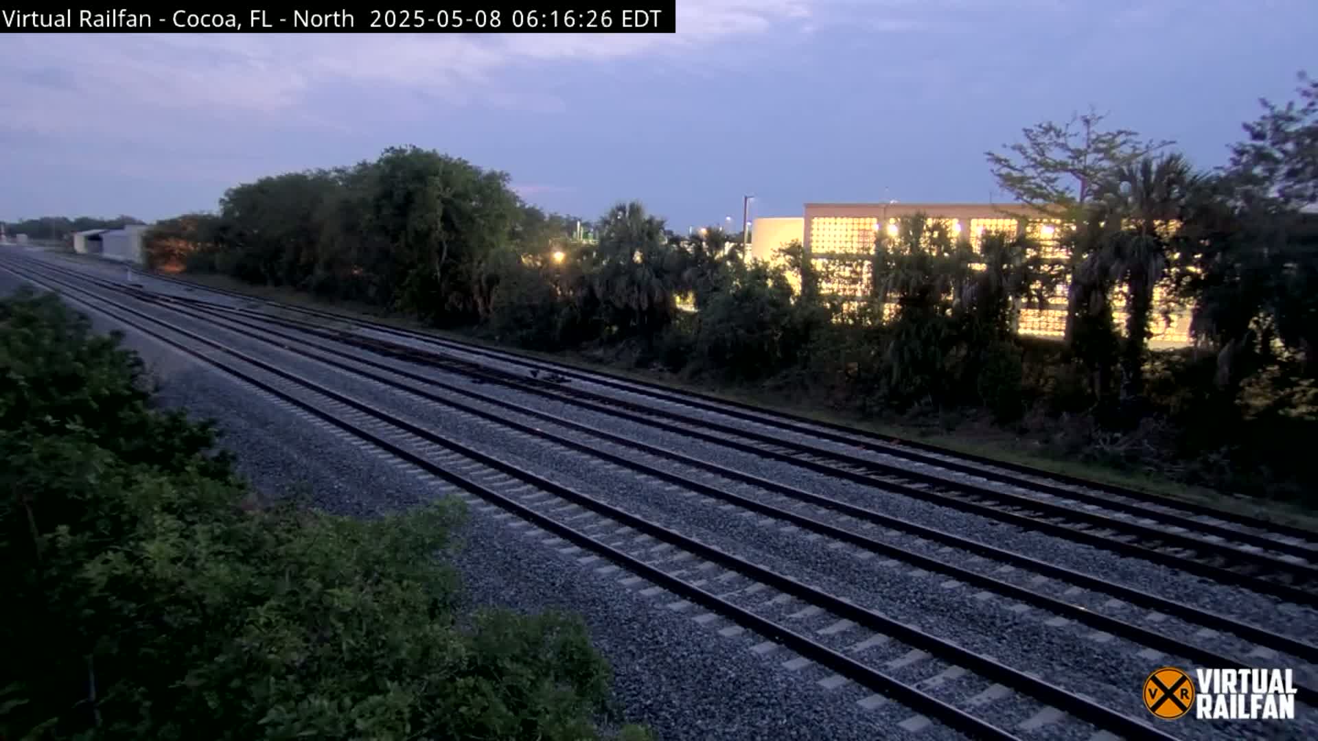 Multiple sets of train tracks stretch into the distance under a twilight sky, passing alongside lush green vegetation and a brightly lit building.