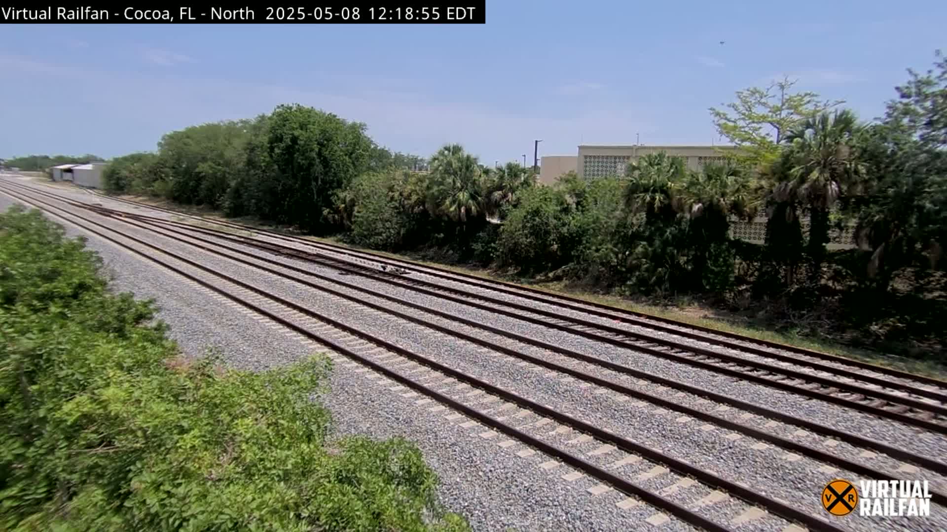 Multiple sets of train tracks stretch into the distance under a clear sky, bordered by lush green vegetation and a building in the background.