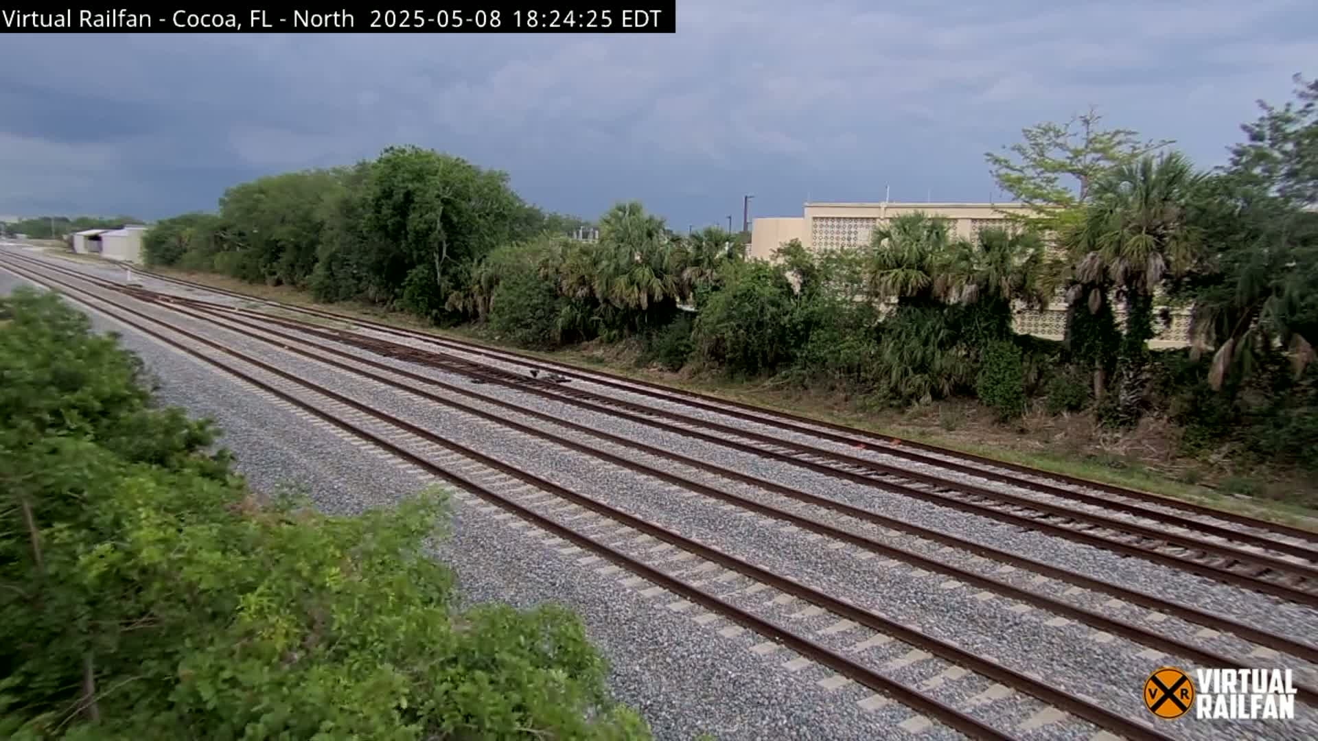 Multiple parallel train tracks extend into the distance under an overcast sky, bordered by lush green vegetation and a building in the background.