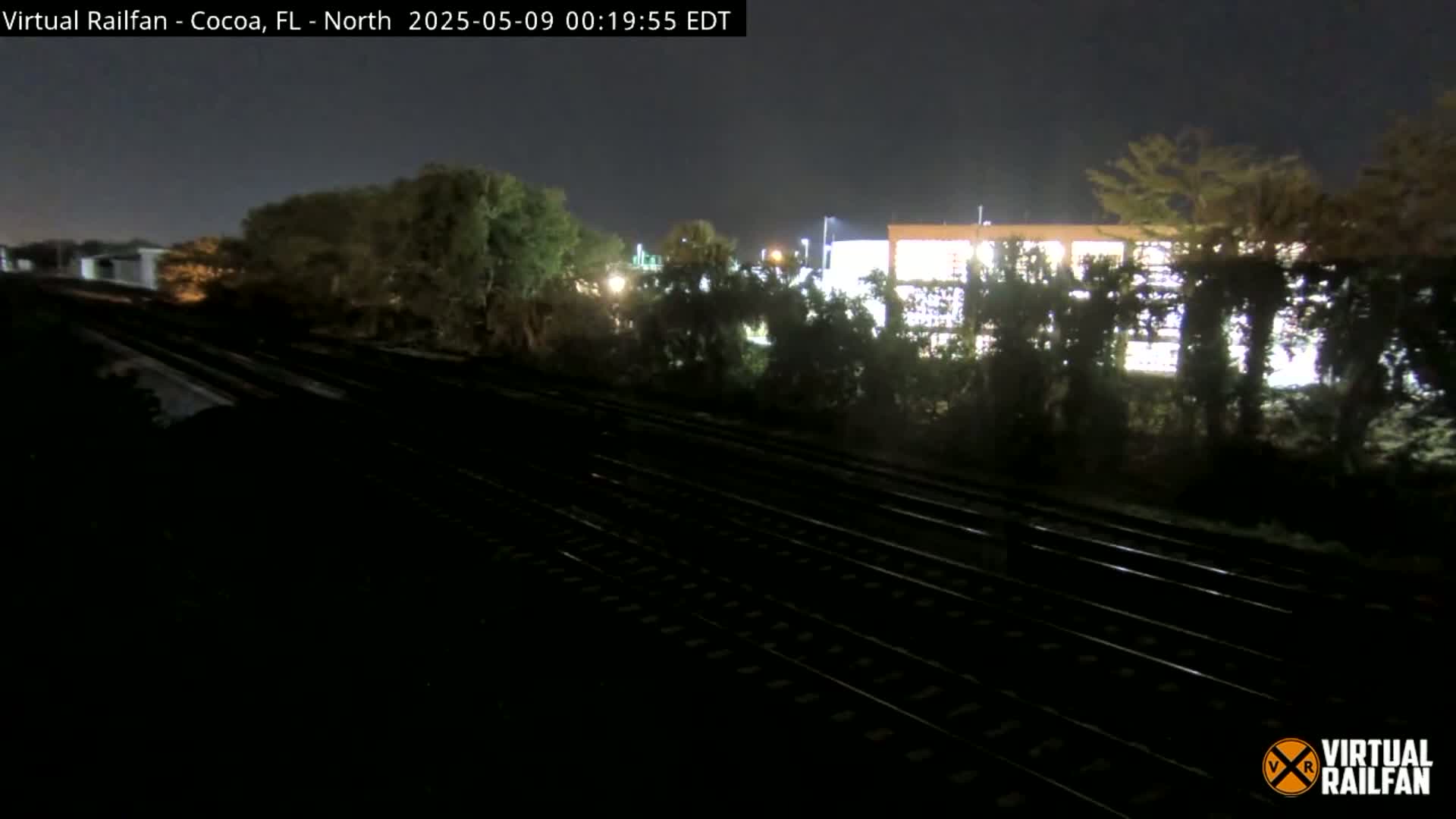 At night, multiple train tracks run past shrubbery and a building, under a dark sky.