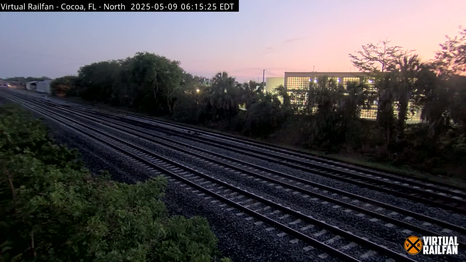 Multiple train tracks stretch into the distance under a dusky, pink-hued sky, bordered by lush green vegetation and a building partially obscured by trees.