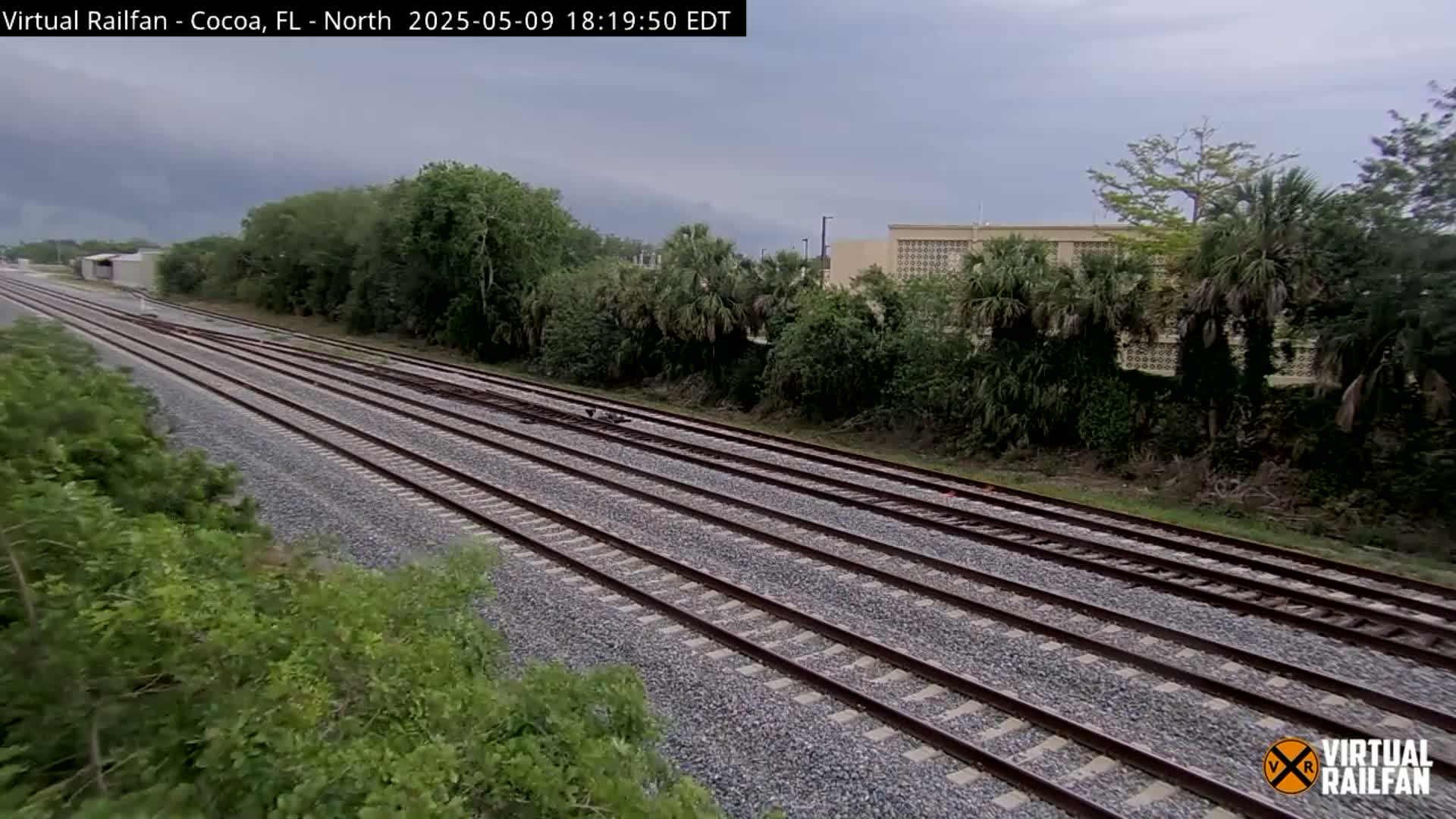 Multiple train tracks stretch into the distance under an overcast sky, bordered by lush green vegetation and a building in the background.