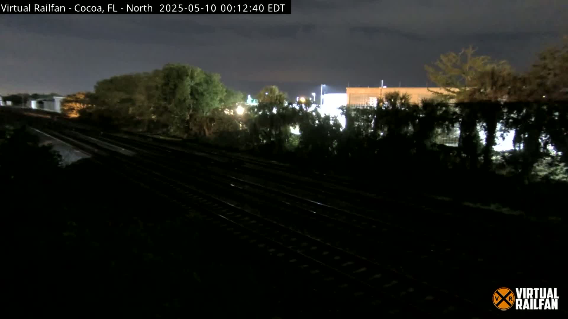 At night, multiple train tracks run through a darkly lit area bordered by trees and buildings under a clear, dark sky.