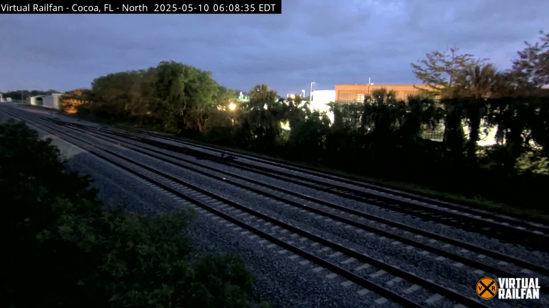 Multiple train tracks stretch into the distance under a dusky sky, bordered by lush green vegetation and buildings.