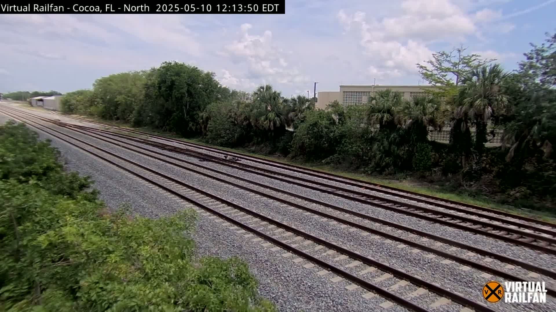Multiple train tracks extend into the distance under a partly cloudy sky, bordered by lush green vegetation and a building.