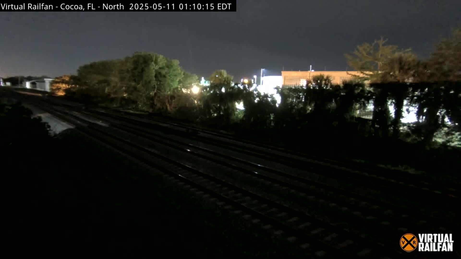 At night, a nighttime view shows multiple parallel train tracks running through a dark landscape with some trees and buildings faintly visible in the background.