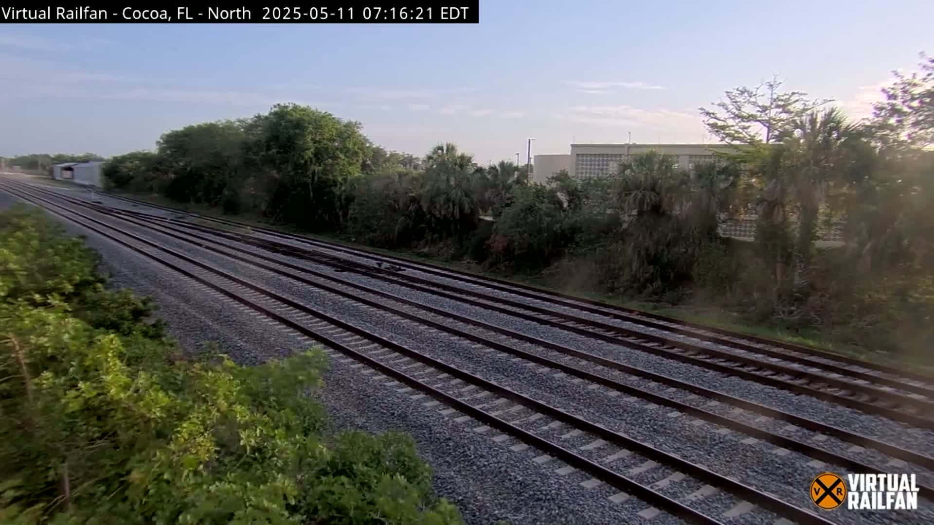 Multiple train tracks stretch into the distance under a partly sunny sky, bordered by lush green vegetation and a building in the background.