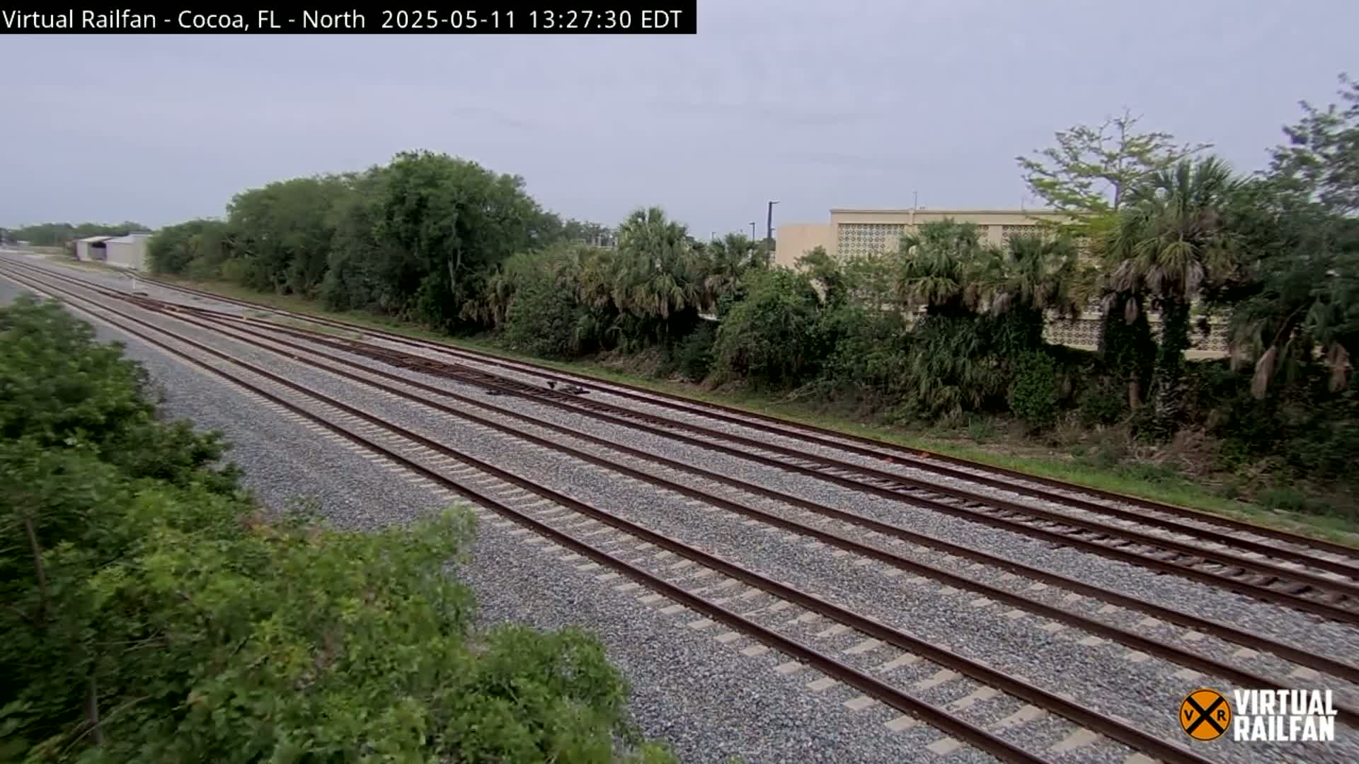 Multiple train tracks stretch into the distance under an overcast sky, bordered by lush green vegetation and a building in the background.