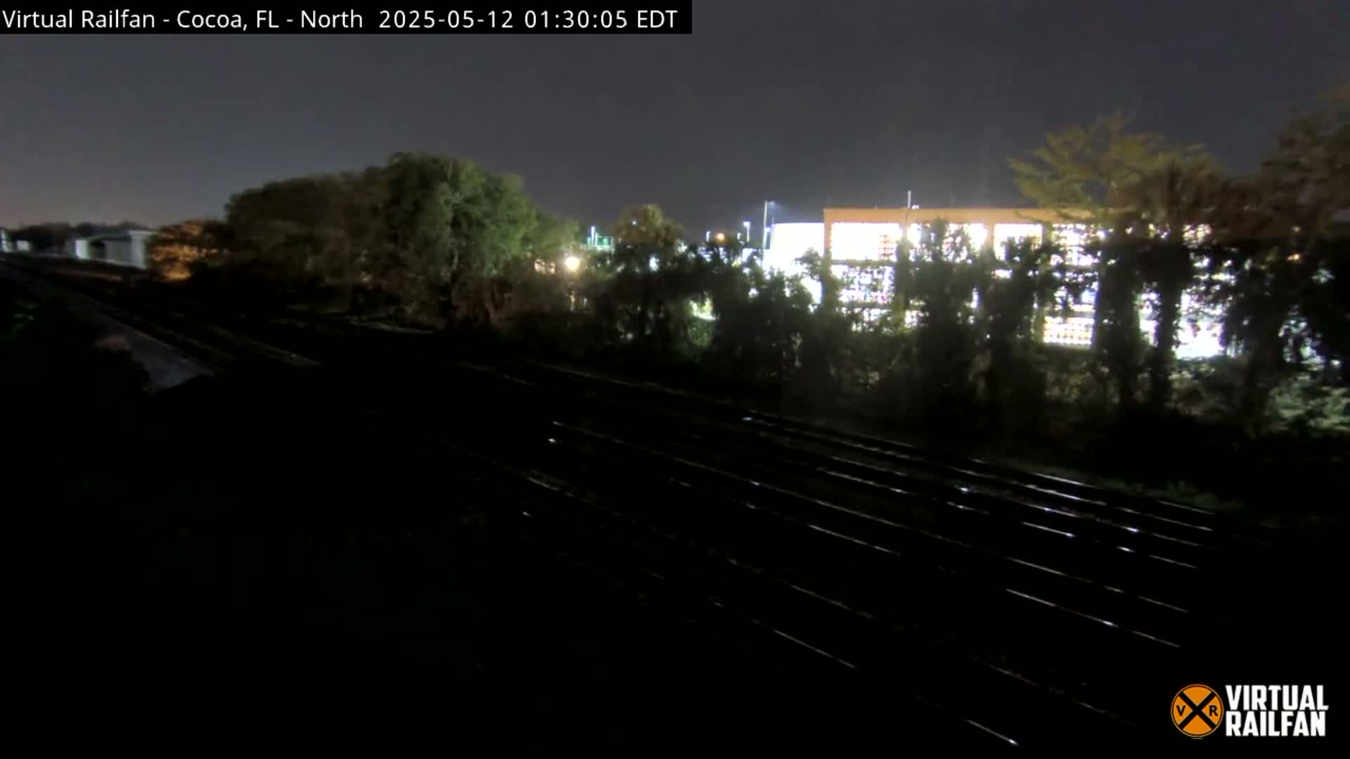 A nighttime view of a railroad track running beside a building partially obscured by trees under a dark sky.