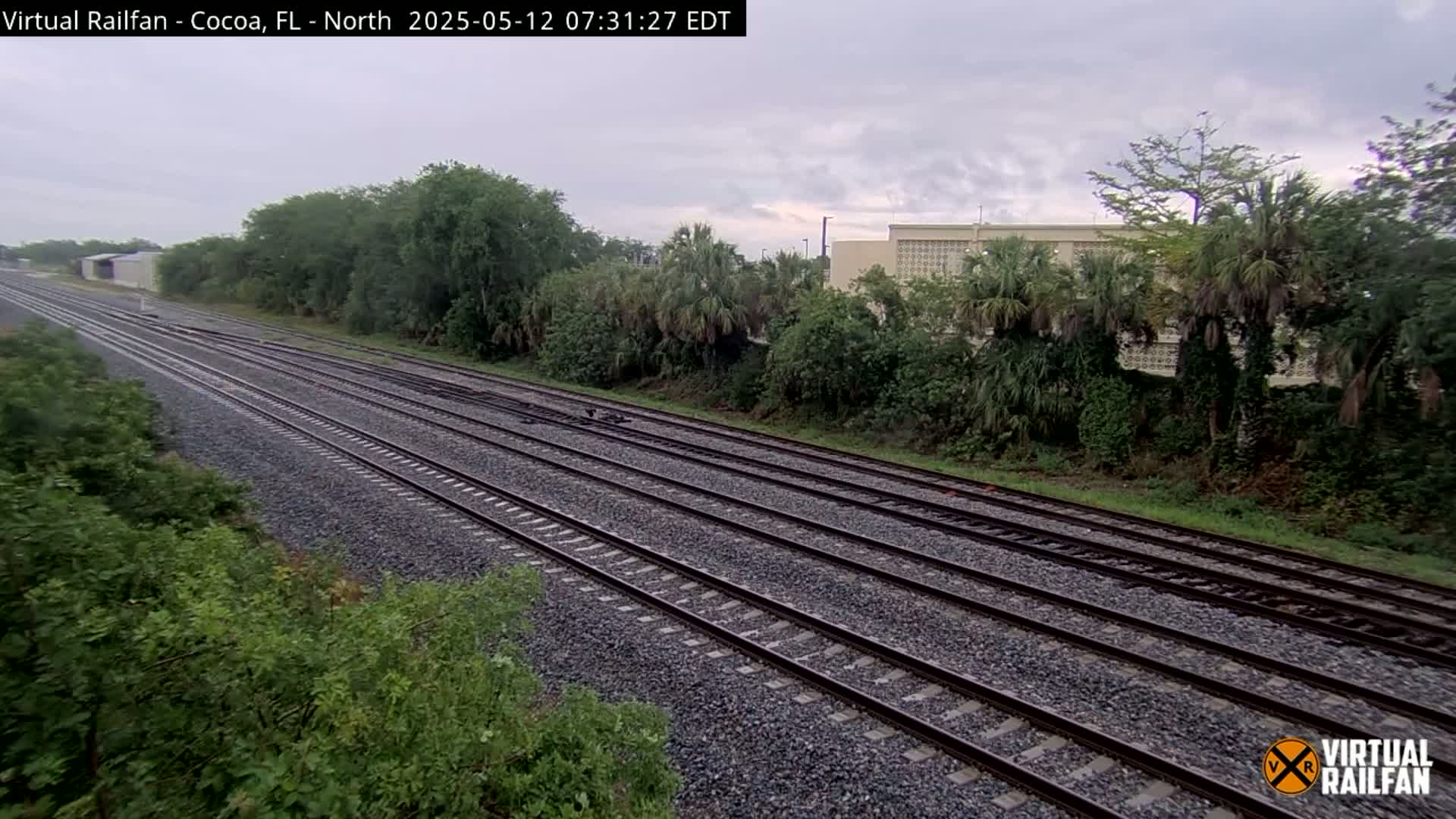 Multiple train tracks stretch into the distance under an overcast sky, bordered by lush green vegetation and a building in the background.