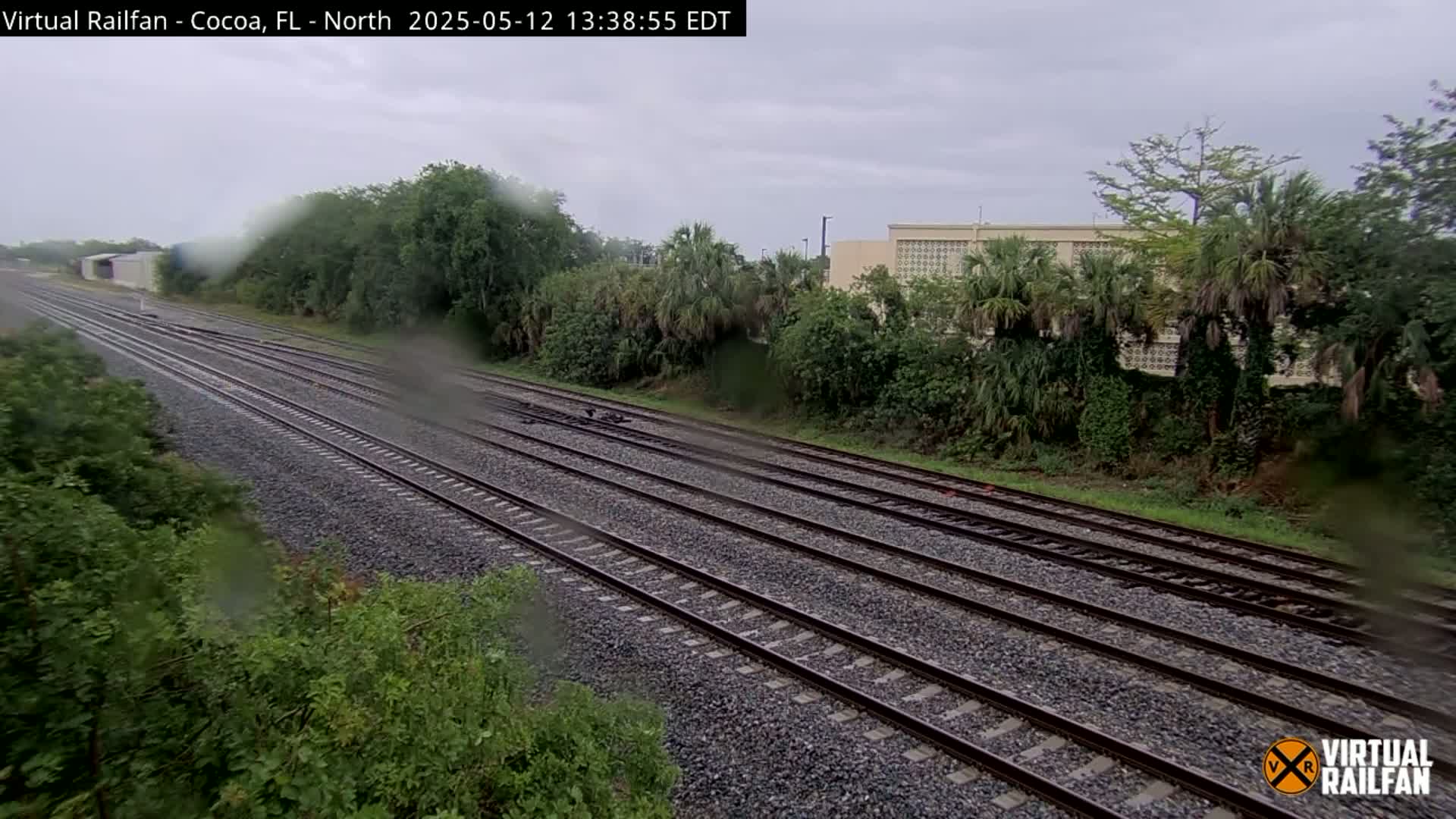 Multiple train tracks stretch into the distance under an overcast sky, surrounded by lush green vegetation and a building in the background, with rain obscuring the view.
