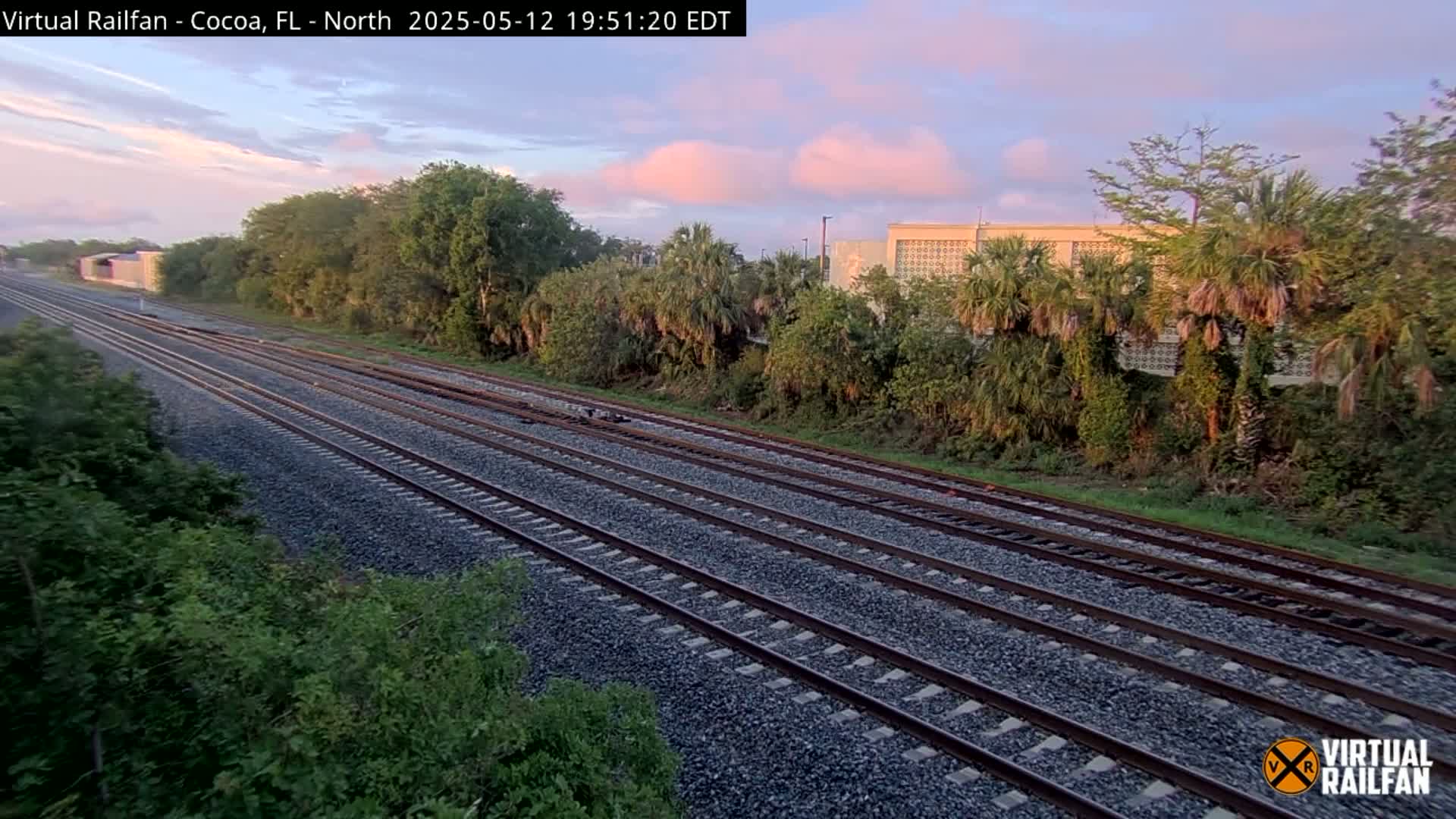 Multiple train tracks extend into the distance, flanked by lush green vegetation under a pale pink and blue sunset sky.
