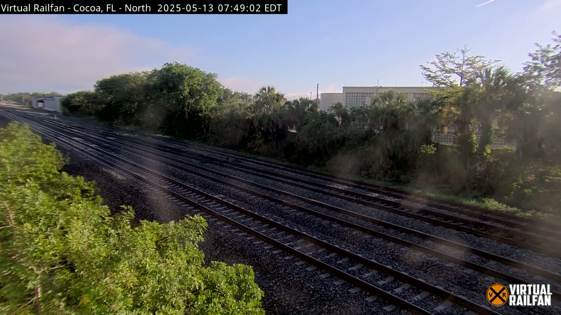 Multiple train tracks stretch into the distance under a clear sky, bordered by lush green vegetation and a building in the background.