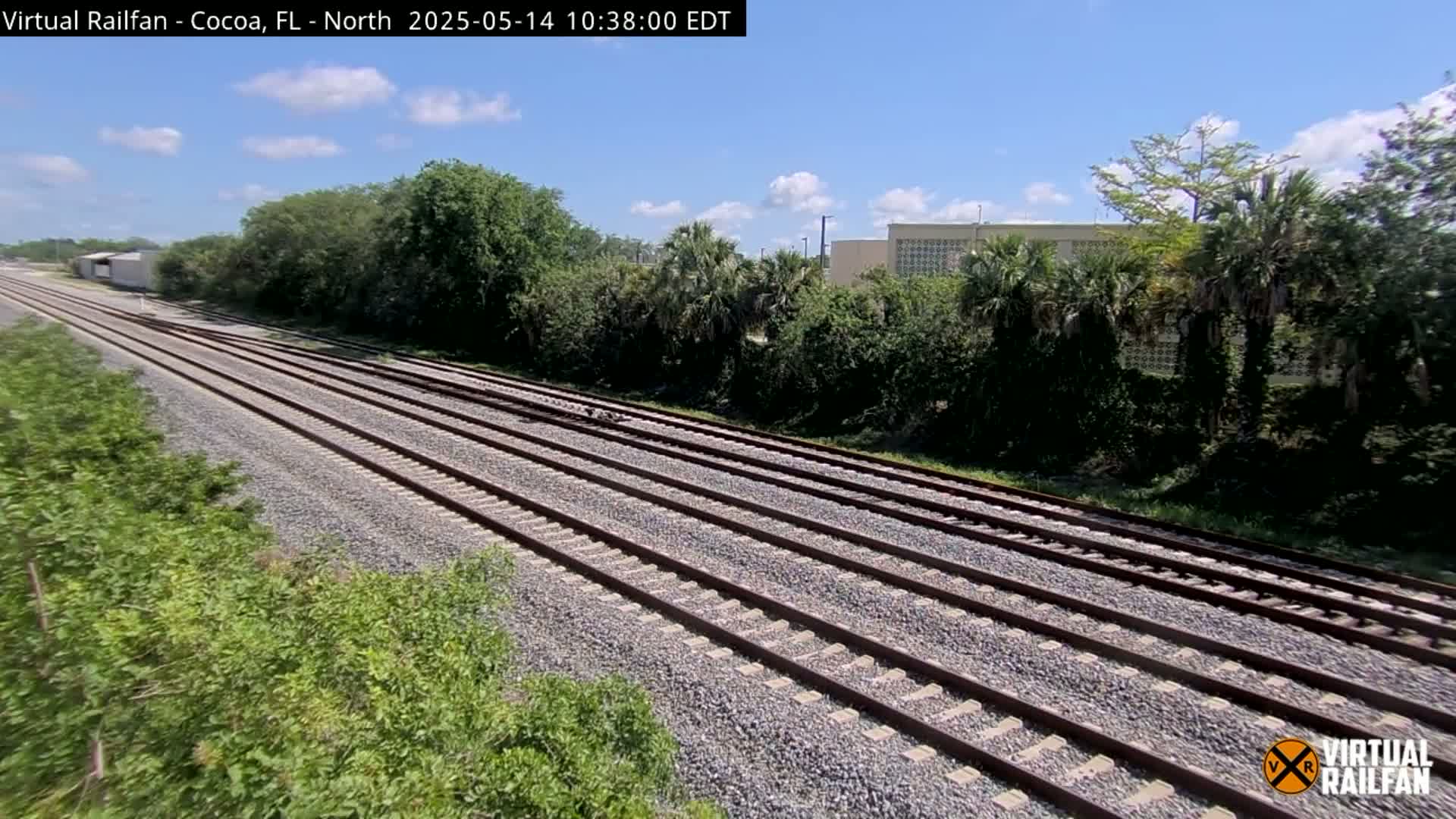Multiple train tracks extend into the distance, bordered by lush green vegetation under a partly cloudy sky.