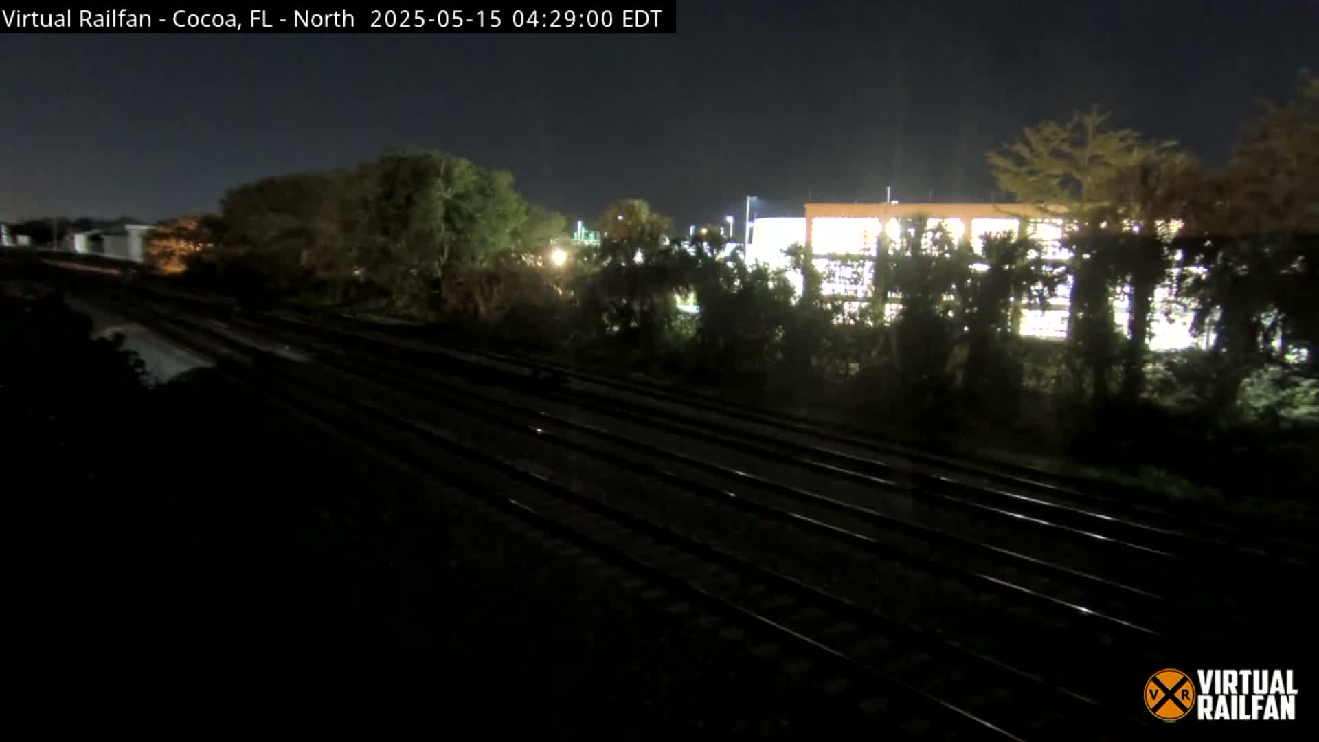 A nighttime view of multiple train tracks running alongside a dark, tree-lined area with a building partially visible in the background.