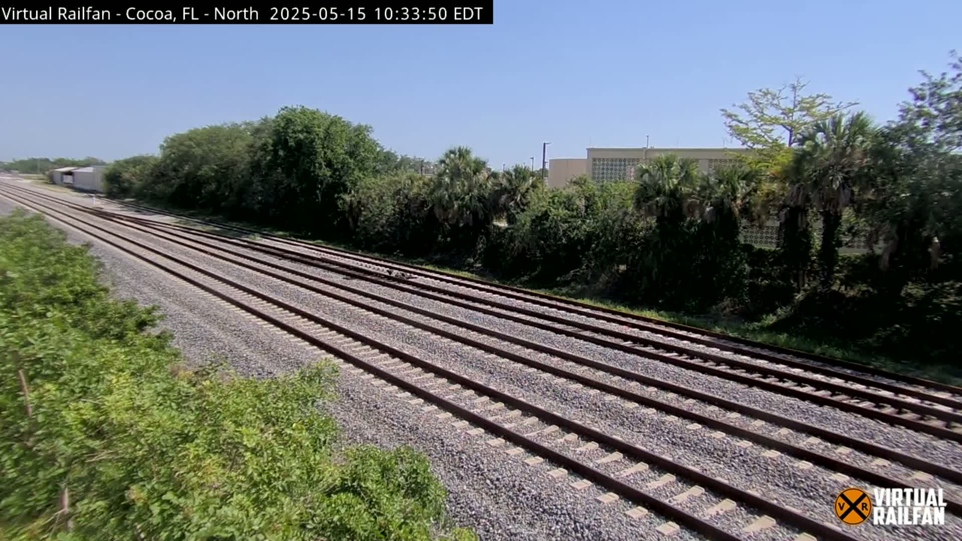Multiple train tracks stretch into the distance under a clear blue sky, flanked by lush green vegetation and a building in the background.