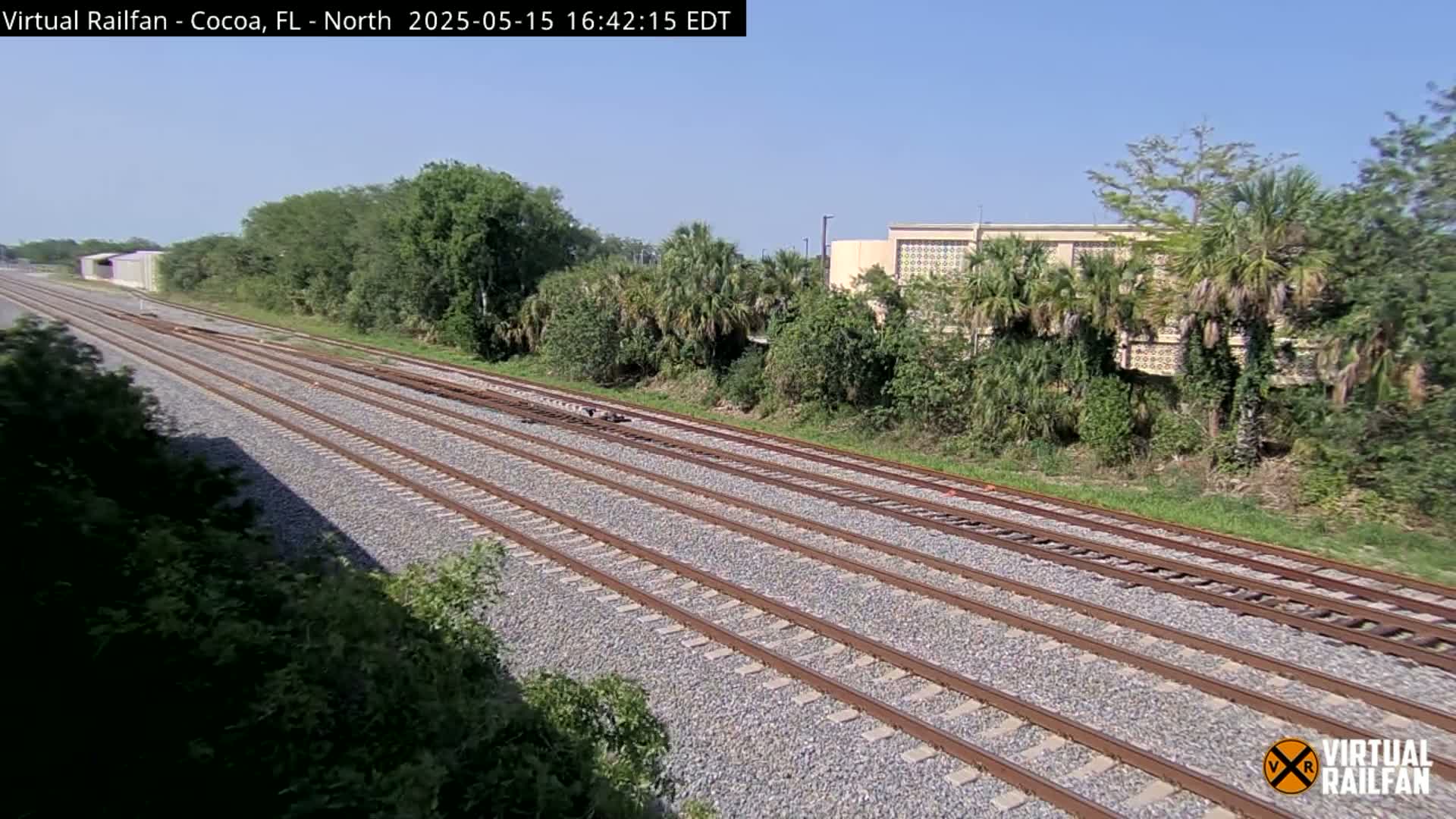 Multiple train tracks stretch into the distance on a sunny day, bordered by lush green vegetation and a building in the mid-ground.