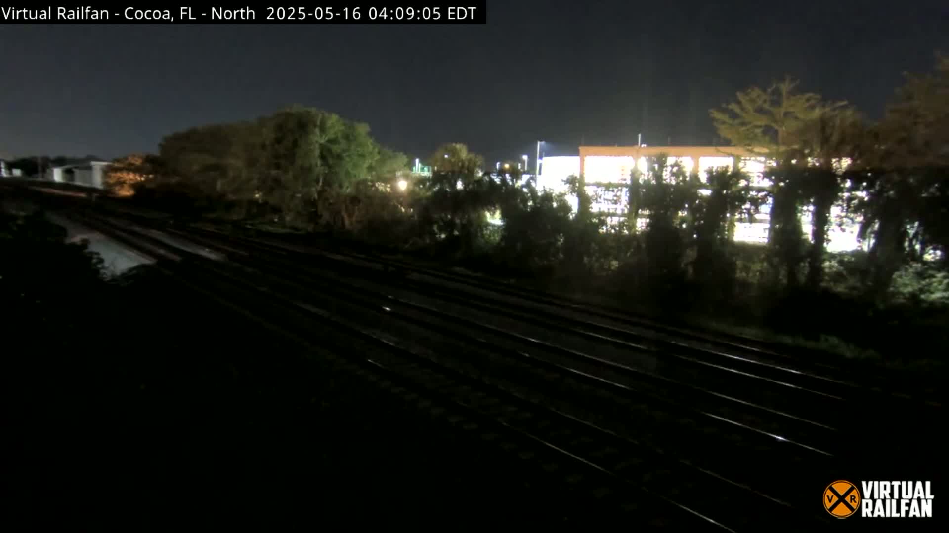 A nighttime view of multiple train tracks running beside a line of trees and a brightly lit building under a dark sky.