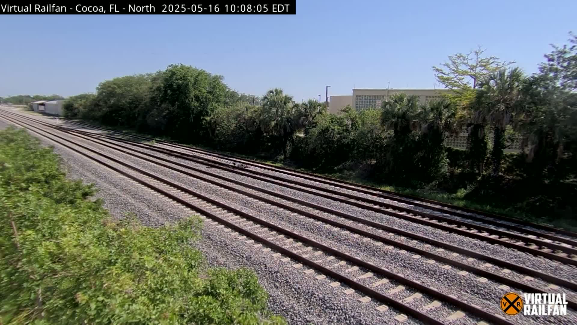 Multiple train tracks extend into the distance on a sunny day, bordered by lush green vegetation and a building in the background.