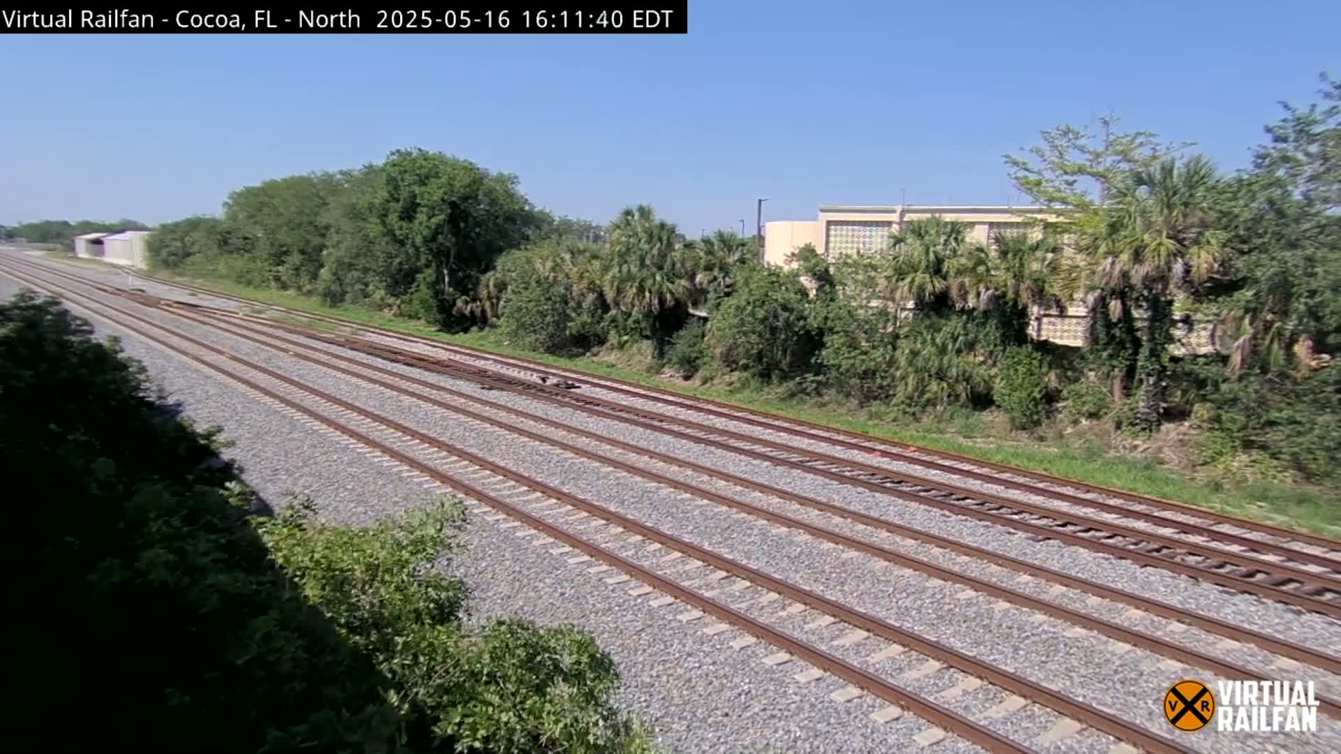 Multiple train tracks extend into the distance on a sunny day, bordered by lush green vegetation and a building in the background.