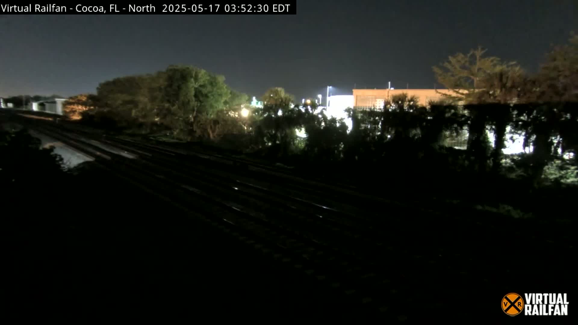 A nighttime view of train tracks running through a dark landscape with some trees and distant lights.