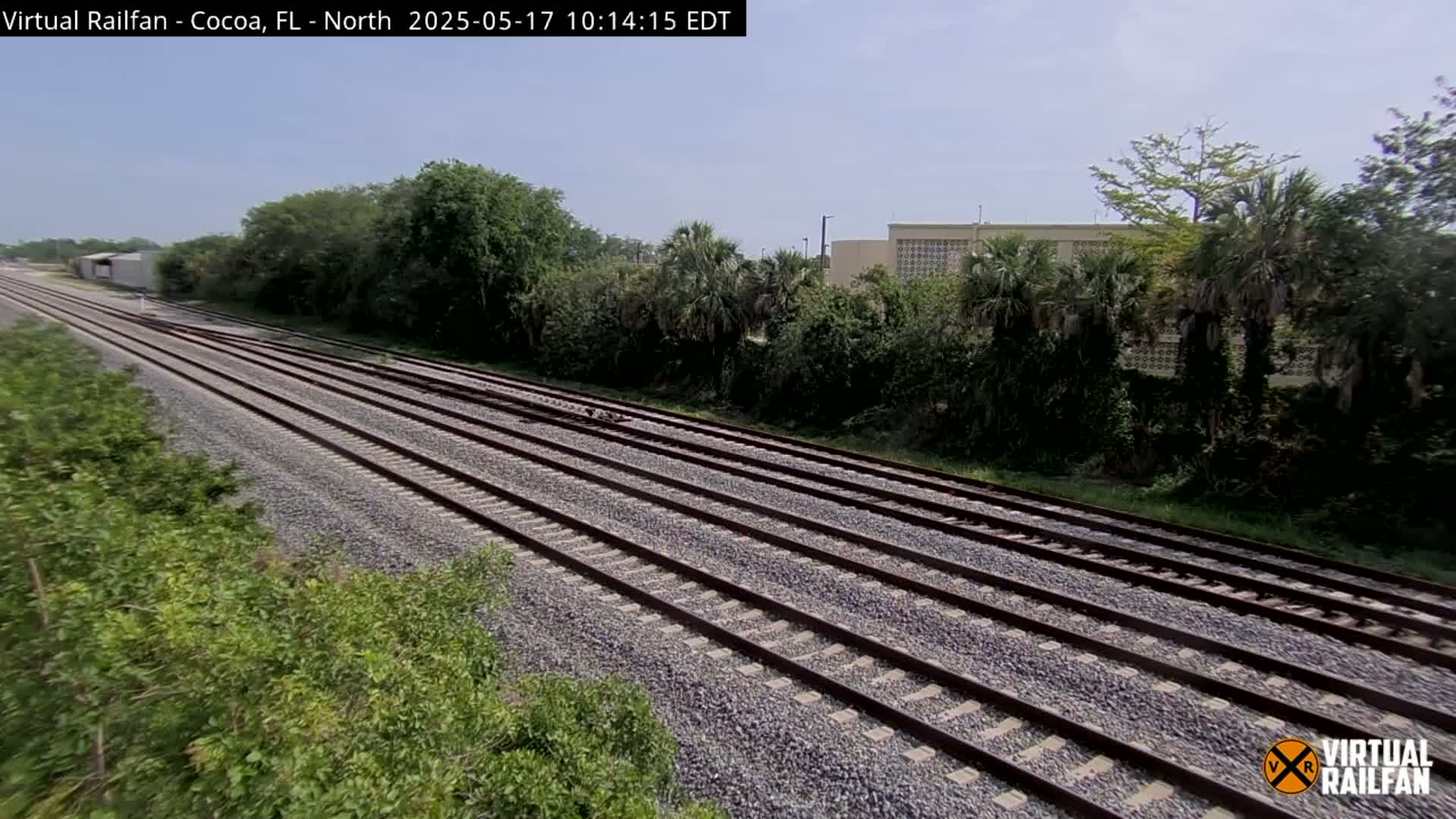 Multiple train tracks extend into the distance under a clear sky, bordered by lush green vegetation and a building in the background.