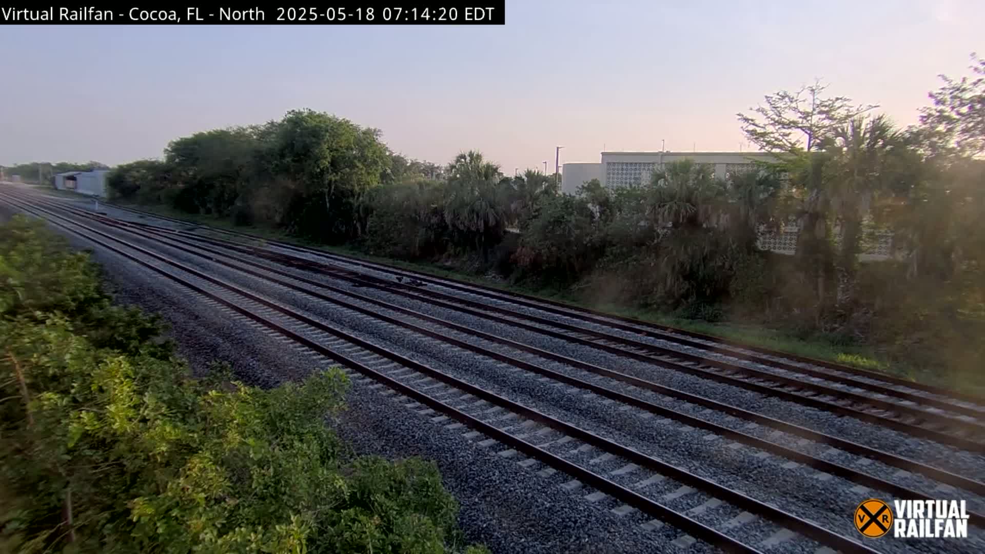 Multiple train tracks extend into the distance under a clear, light-colored sky, bordered by lush green vegetation and a building in the background.