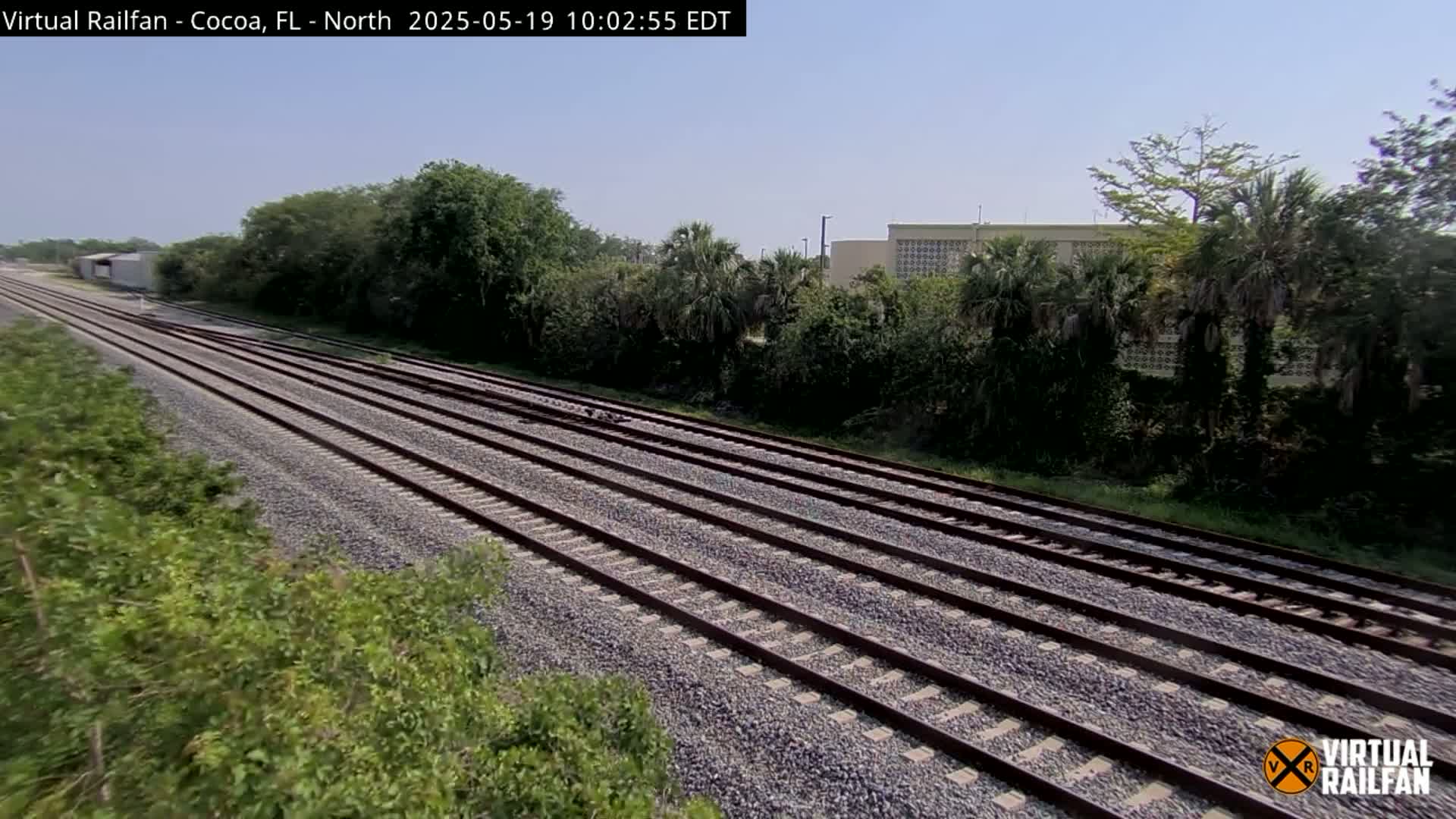 Multiple parallel train tracks stretch into the distance under a clear sky, bordered by lush green vegetation and a building in the background.