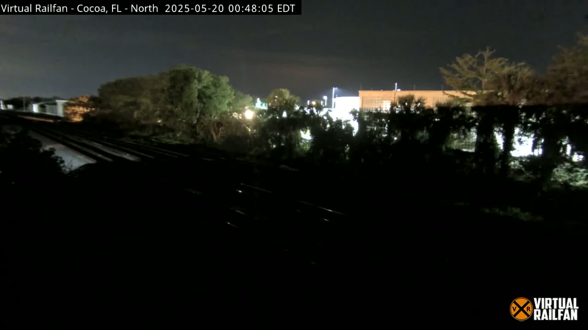 A nighttime view of train tracks running through a wooded area next to a dimly lit building under a clear sky.