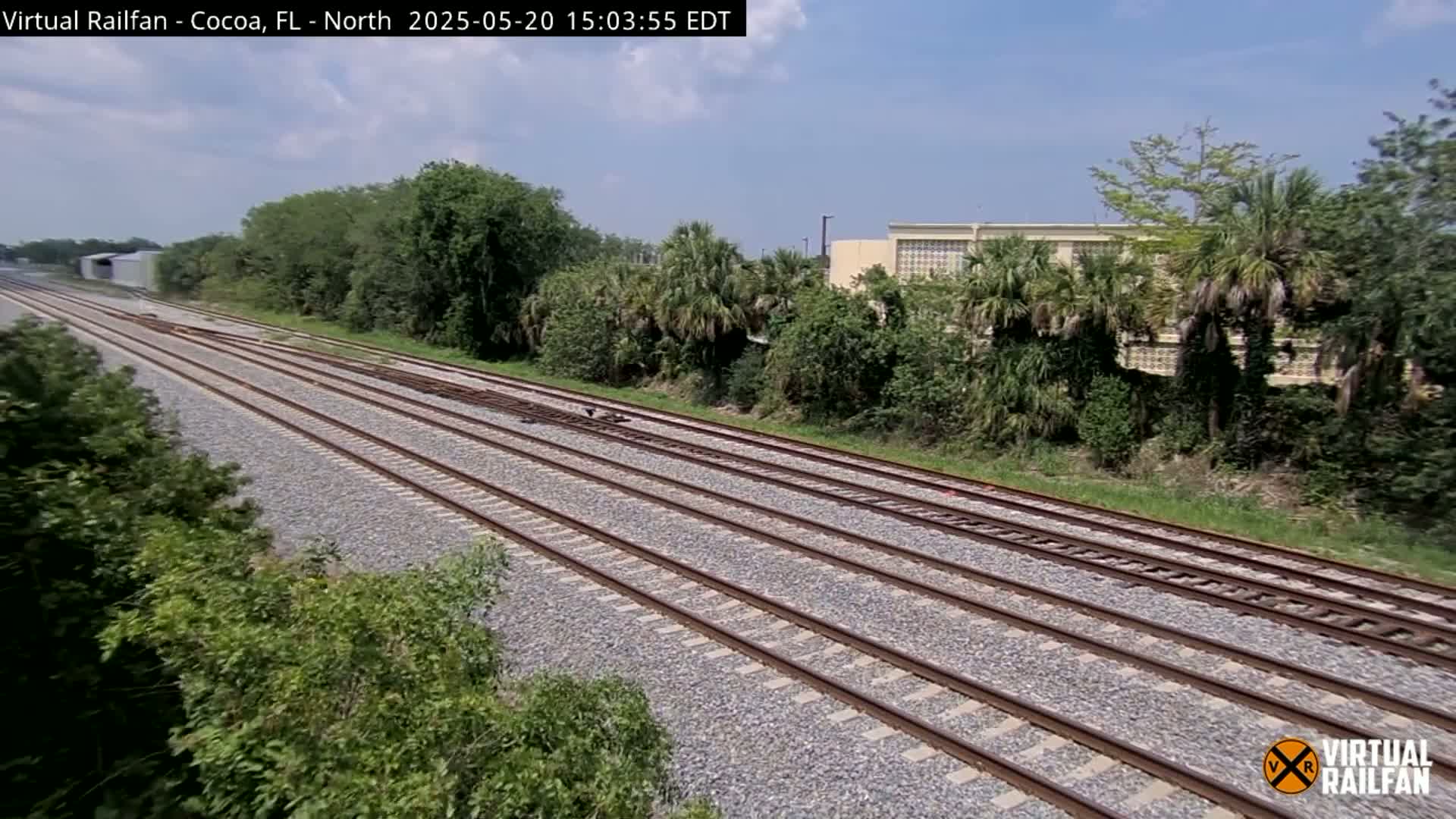 Multiple train tracks extend into the distance under a sunny sky, bordered by lush green vegetation and a building in the background.