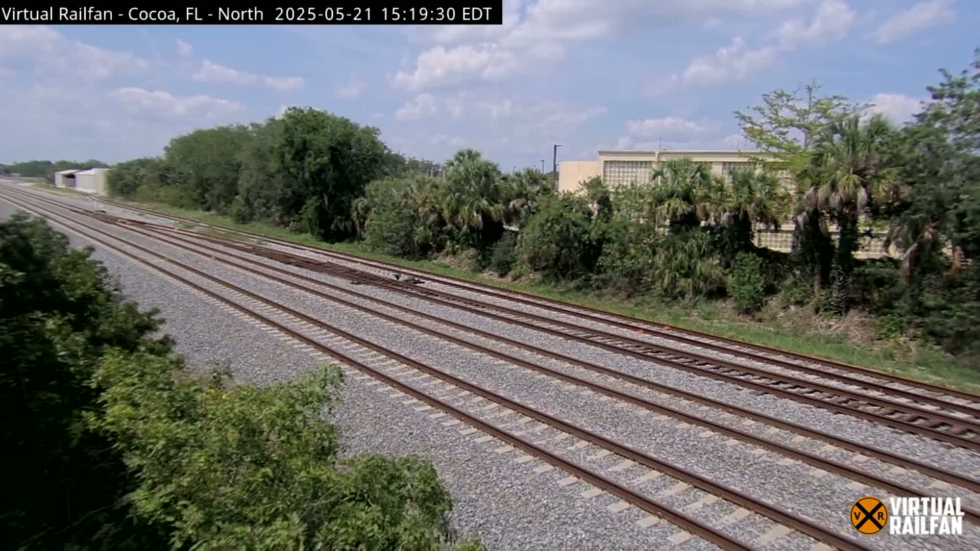 Multiple train tracks stretch into the distance under a partly cloudy sky, bordered by lush green vegetation and a building in the background.