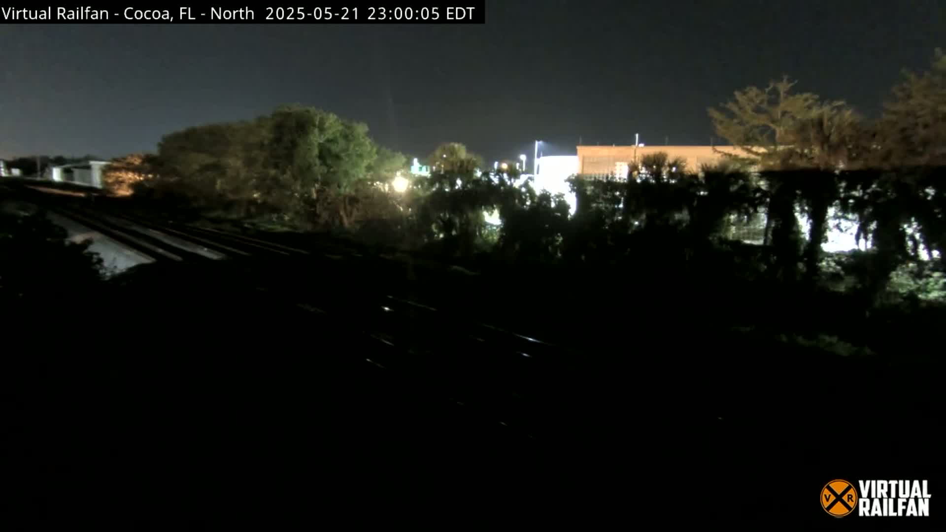 At night, train tracks run through a dark landscape of trees and dimly lit buildings under a clear sky.