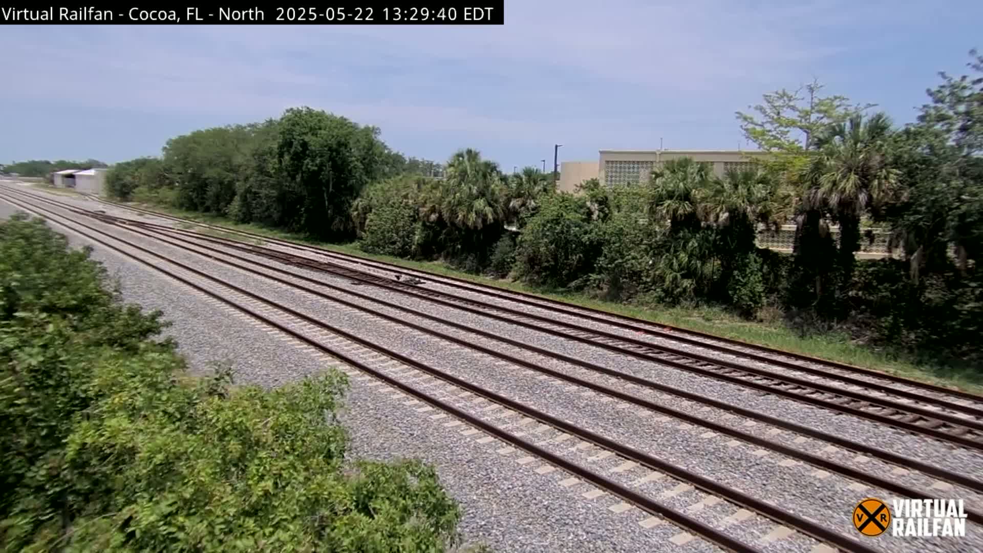 Multiple train tracks extend into the distance under a partly sunny sky, bordered by lush green vegetation and a building in the background.