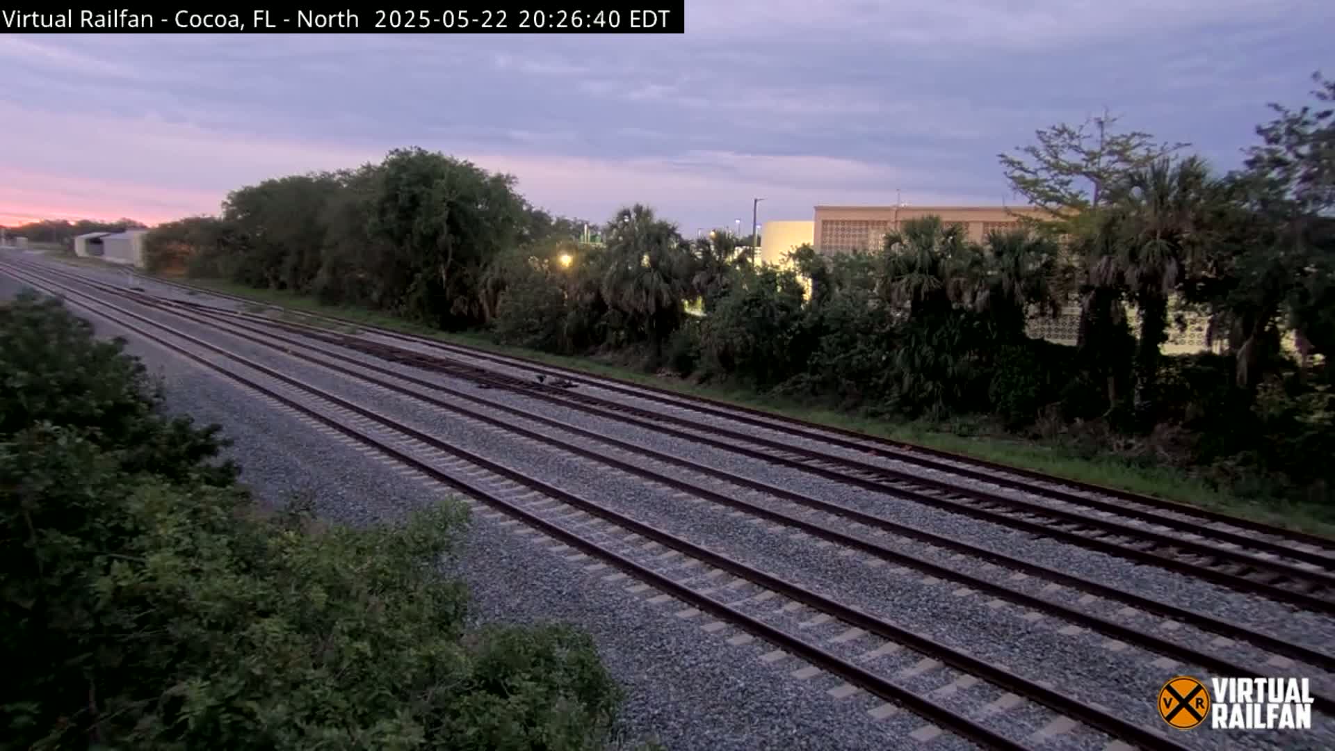 Multiple train tracks extend into the distance, bordered by lush green vegetation under a dusky, twilight sky.