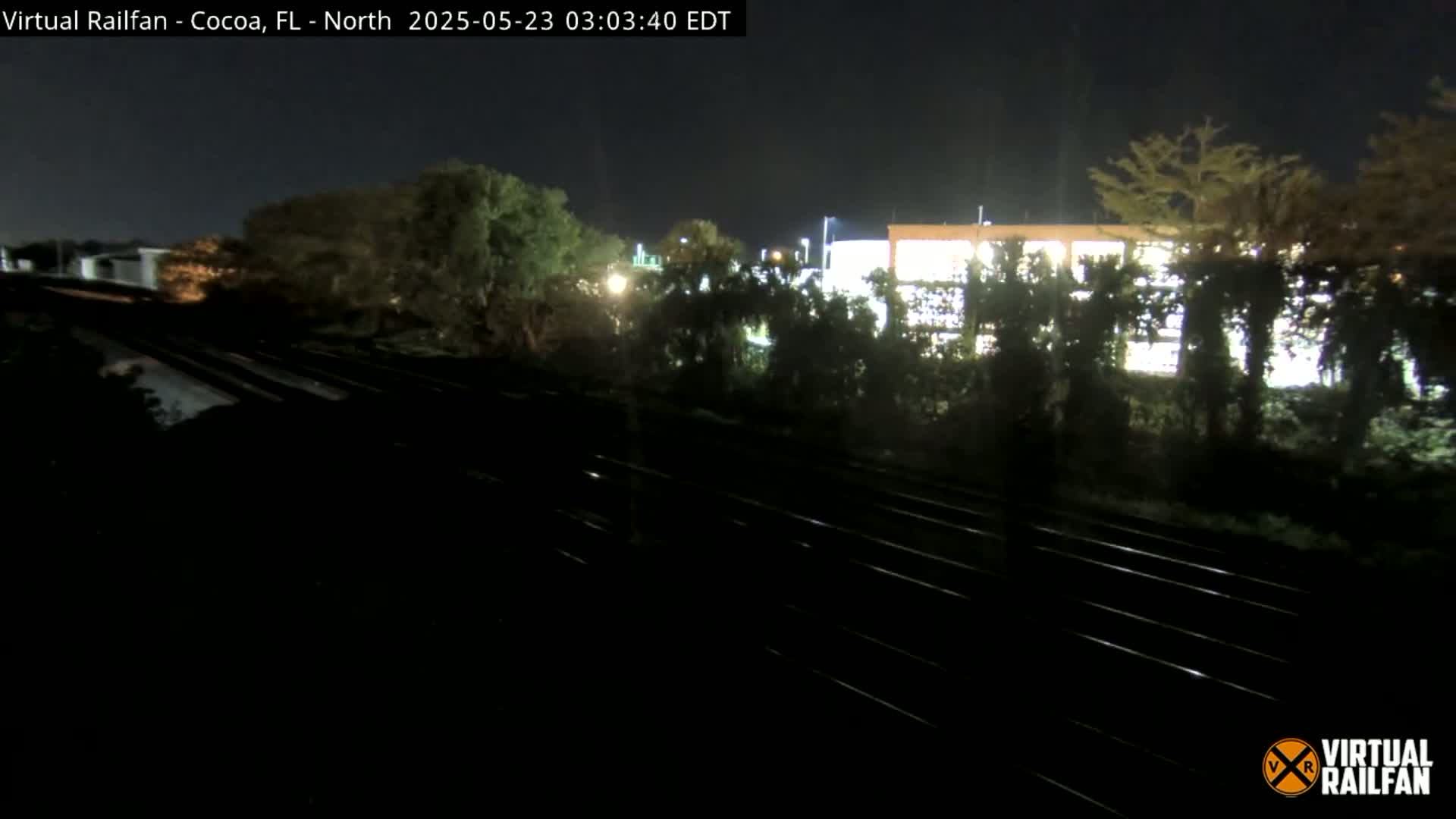 At night, a nighttime view shows train tracks in front of a partially visible, brightly lit building behind a line of trees.