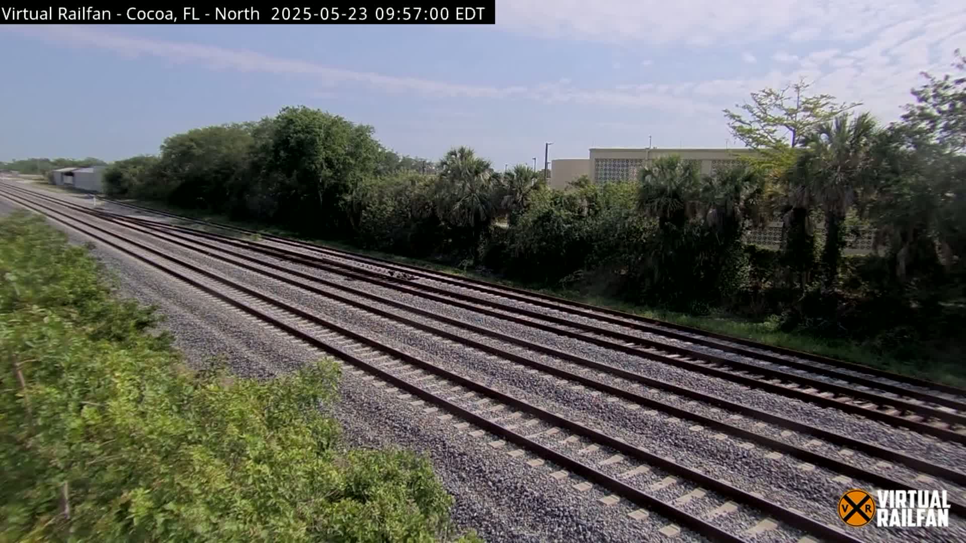 Multiple sets of train tracks stretch into the distance, bordered by lush green vegetation under a partly sunny sky.